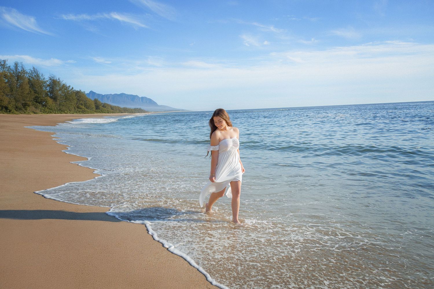 Person in flowing white dress walks along pristine Hawaiian beach shoreline with turquoise waters and mountains in distance.