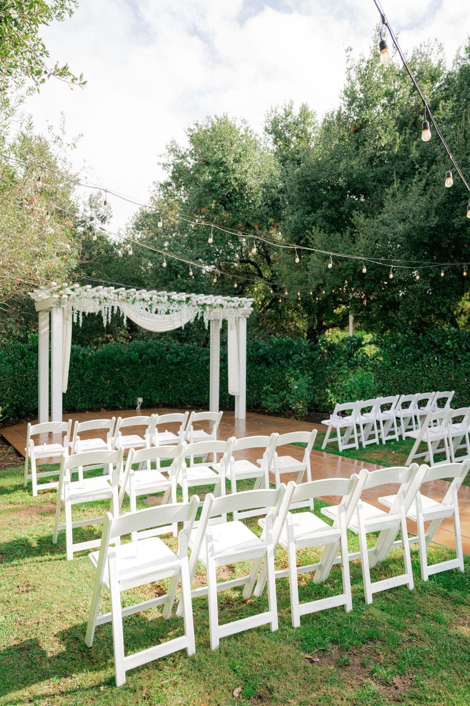 White folding chairs arranged in rows on a grassy lawn beneath string lights and a white wedding arch for an outdoor ceremony.