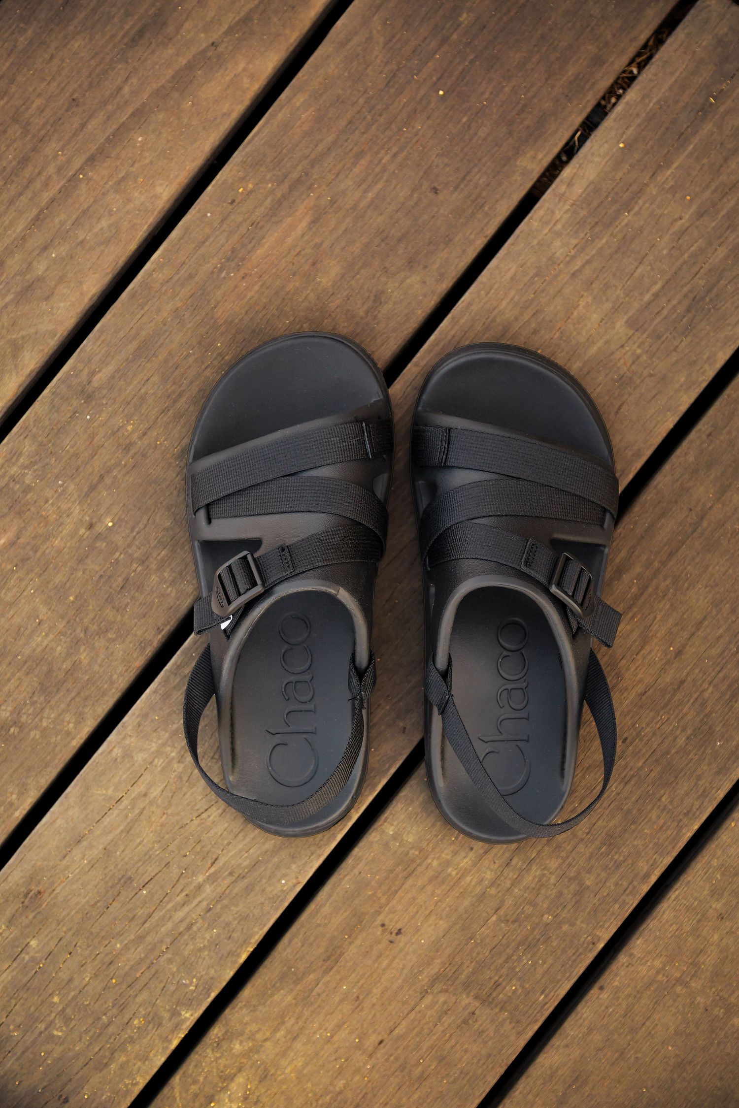 Black leather Birkenstock-style sandals with adjustable straps shown from different angles on wooden deck planks.