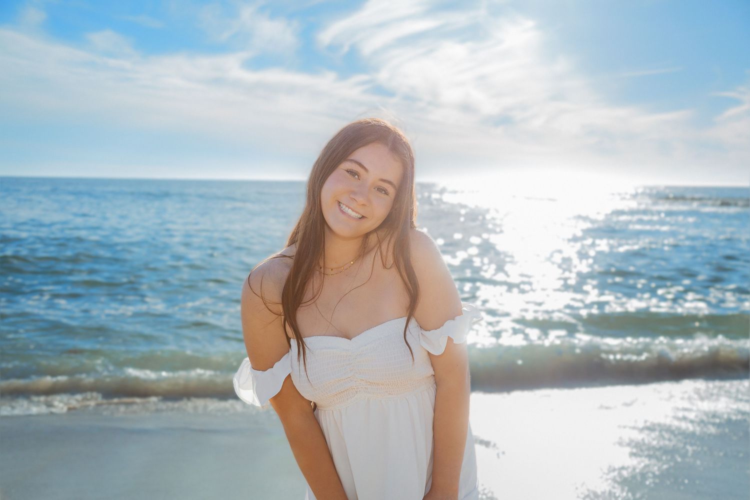 Person in a white dress poses on a sunny beach with sparkling ocean waves and bright sunlight in the background.