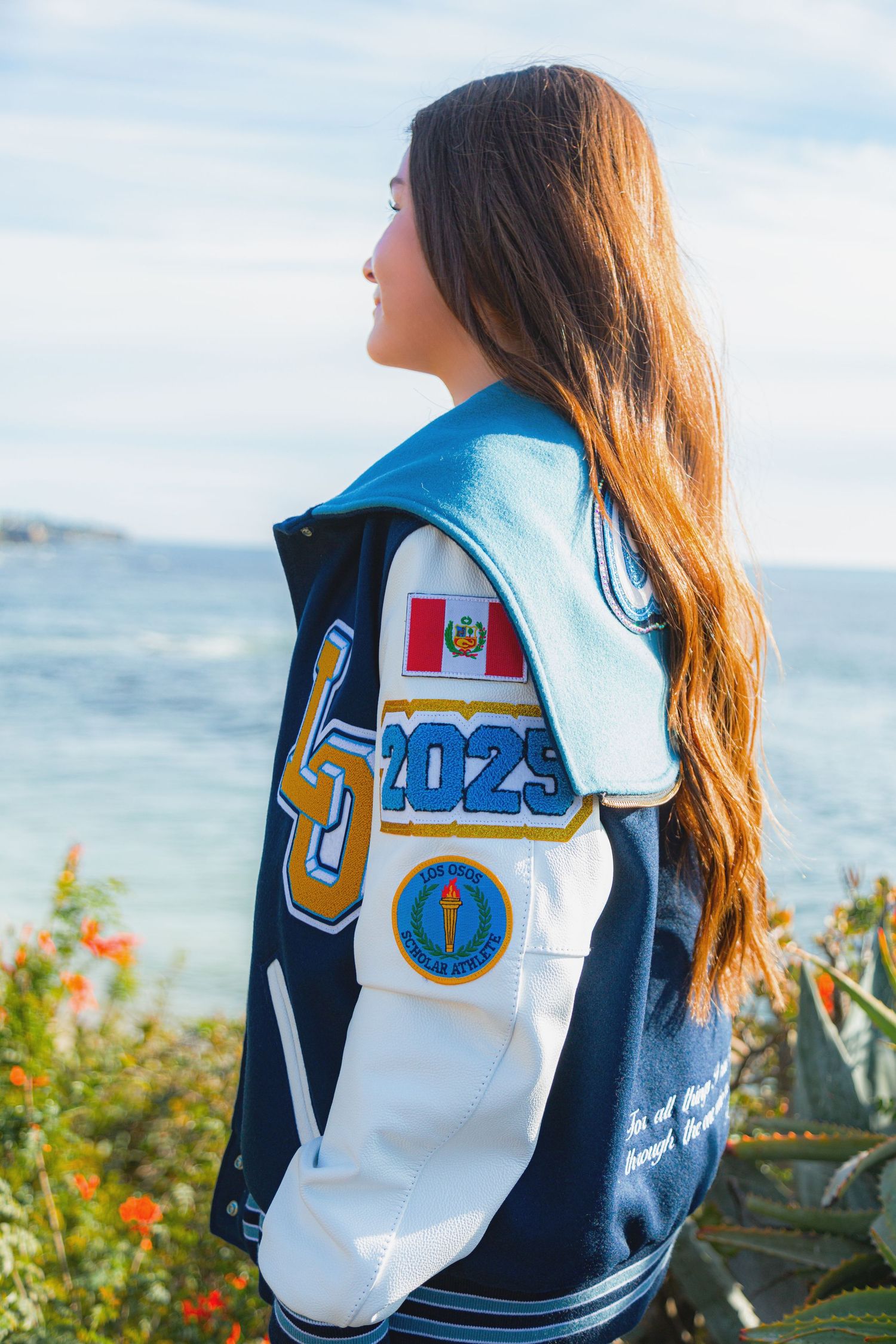 A person wearing a navy and white varsity letterman jacket poses against an ocean backdrop with flowers in the foreground.
