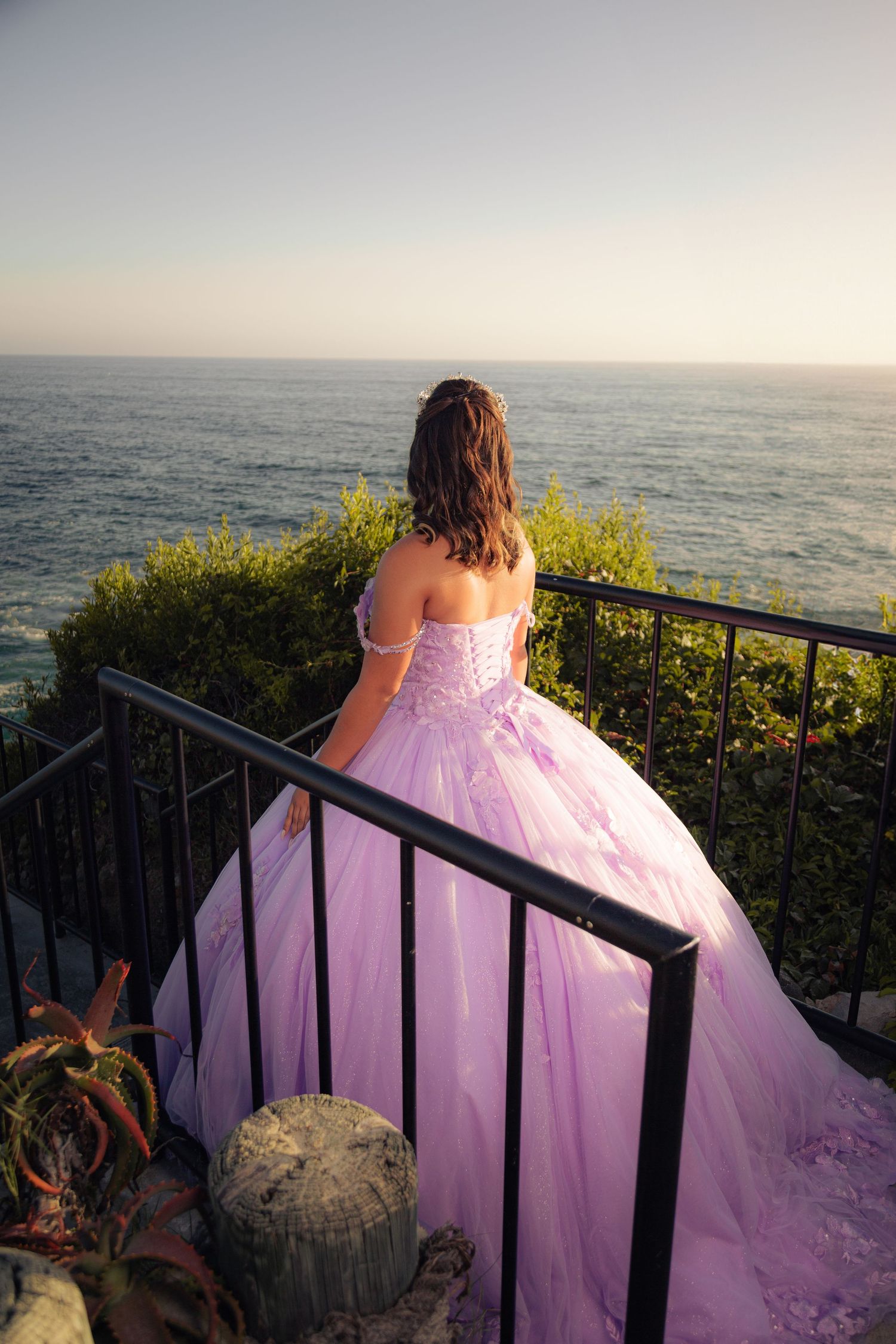 Someone in a flowing pink gown stands at an oceanside railing during sunset, creating a romantic silhouette.