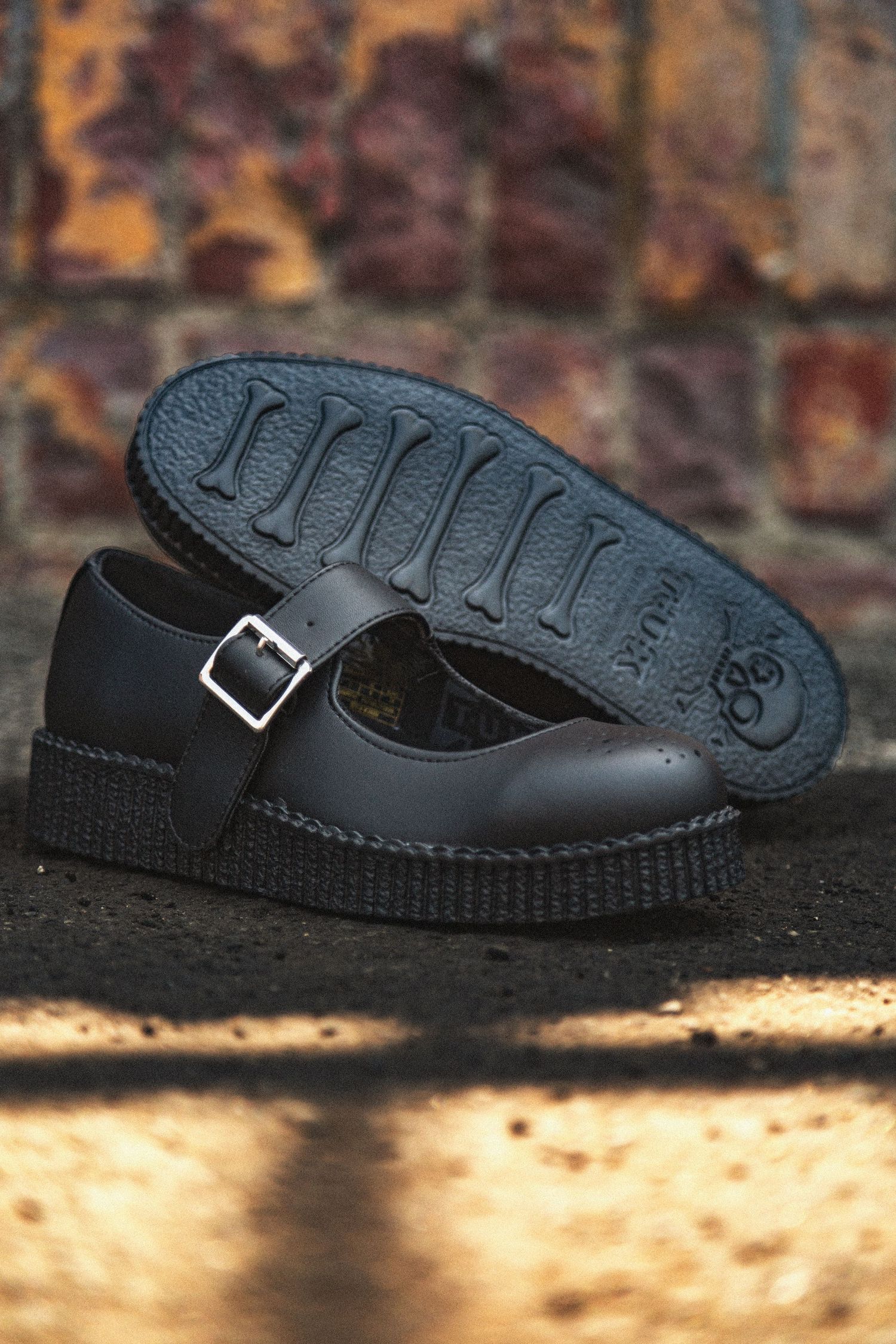 Black leather Mary Jane creeper shoe with buckle strap and thick platform sole against brick wall background.