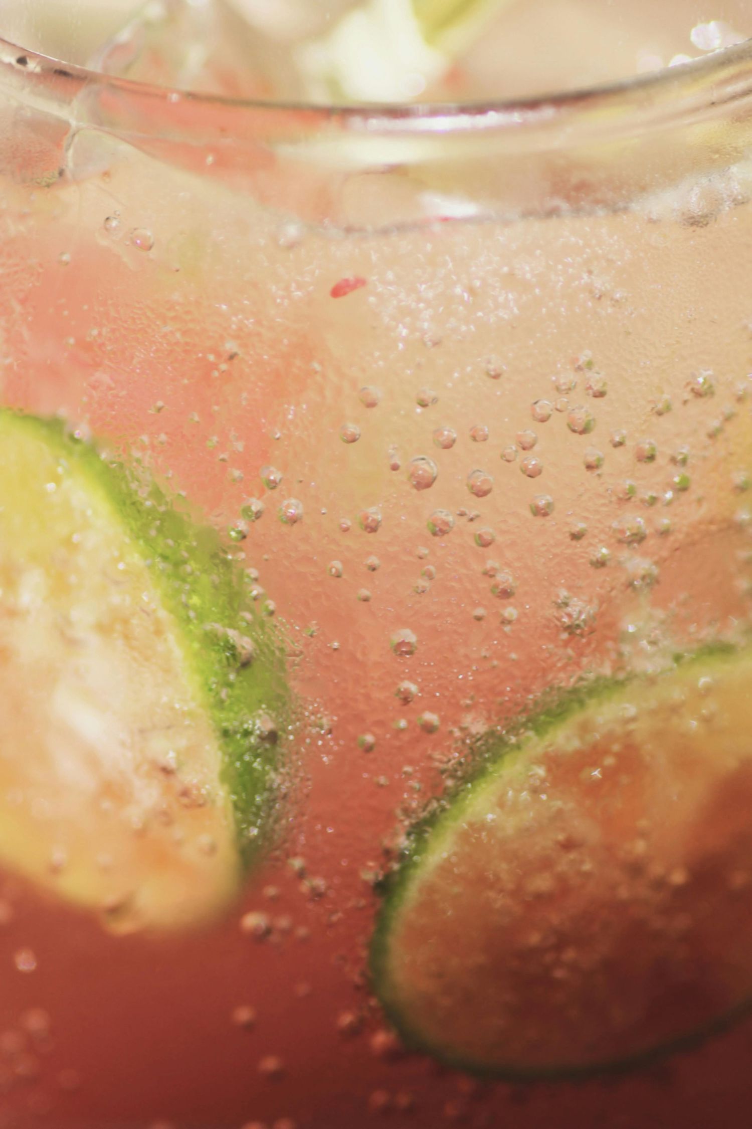Close-up macro shot of carbonated beverage with lime slices and bubbles in pink-tinted liquid.