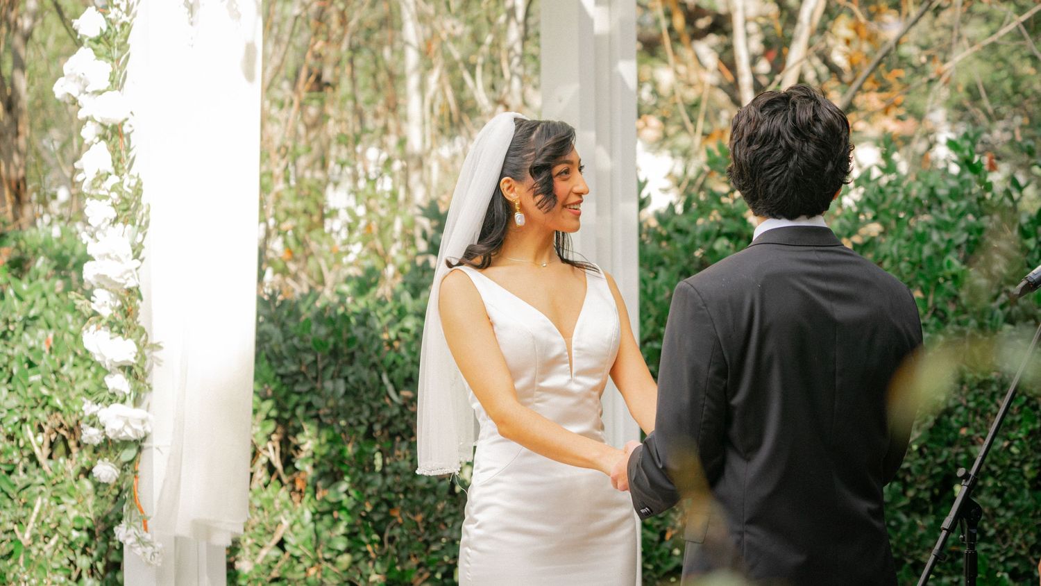 A wedding couple walks through a lush garden path surrounded by white flowering trees and greenery.