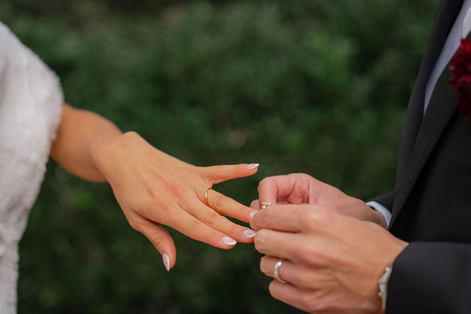 A close-up shot of hands exchanging wedding rings during an outdoor ceremony against a green background.