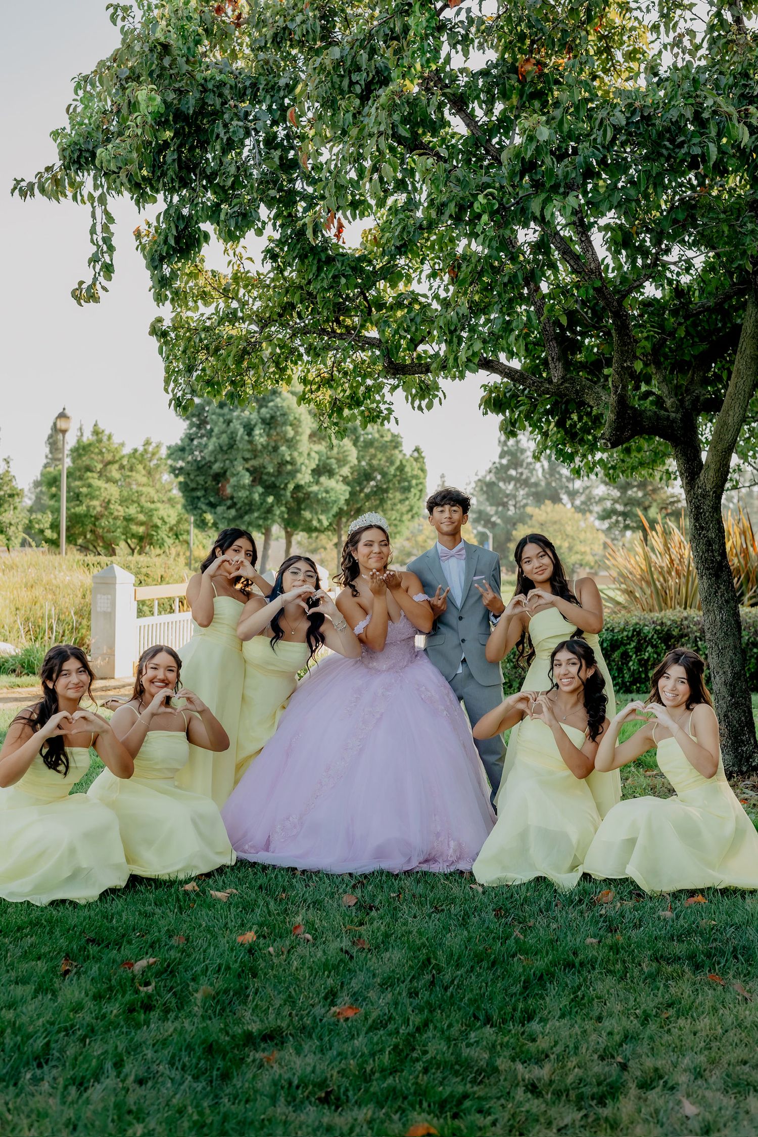 A bridal party in yellow and purple dresses poses playfully on a grassy hill under trees.