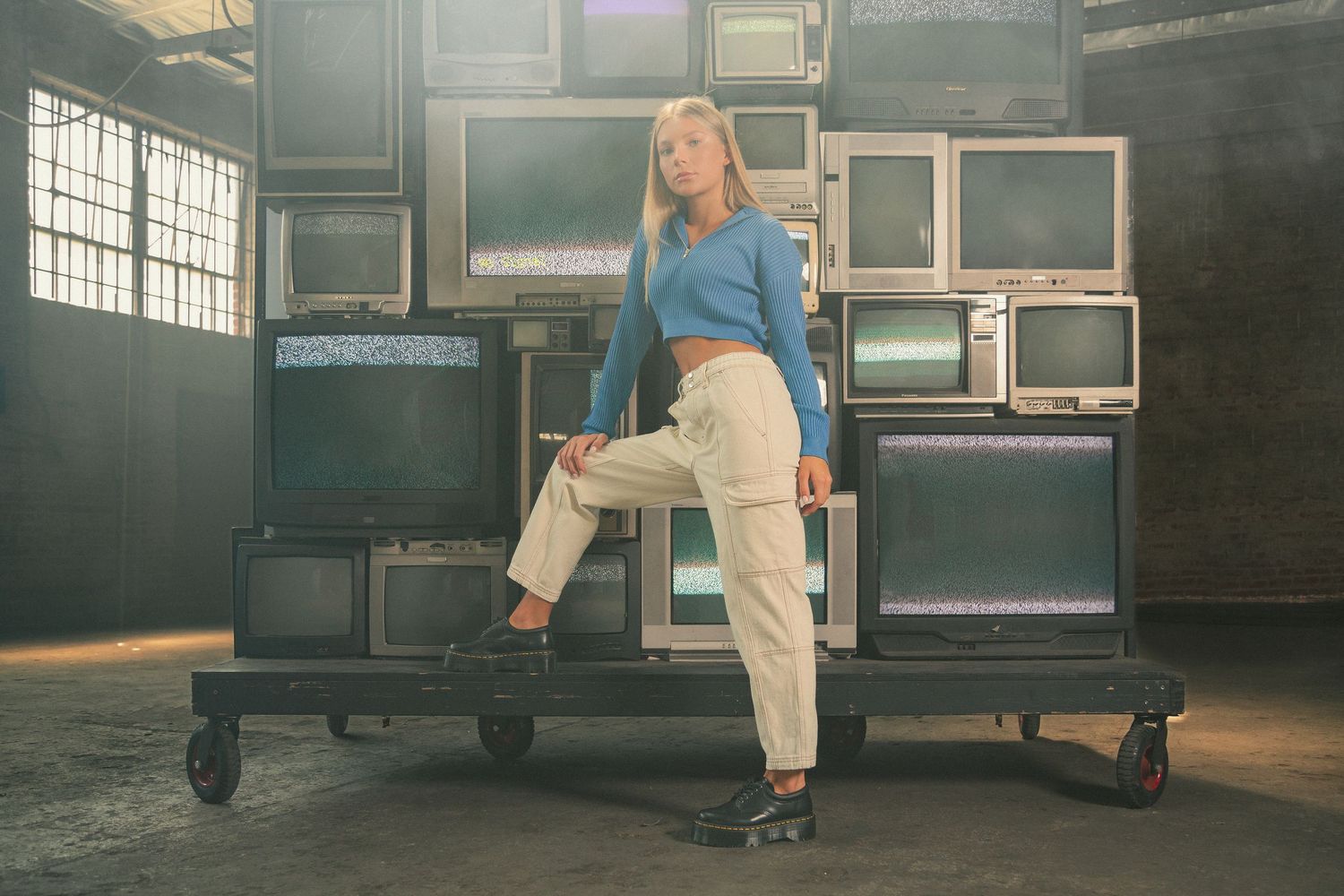 A dancer in white pants and a blue shirt performs graceful movements in a sunlit industrial studio space.