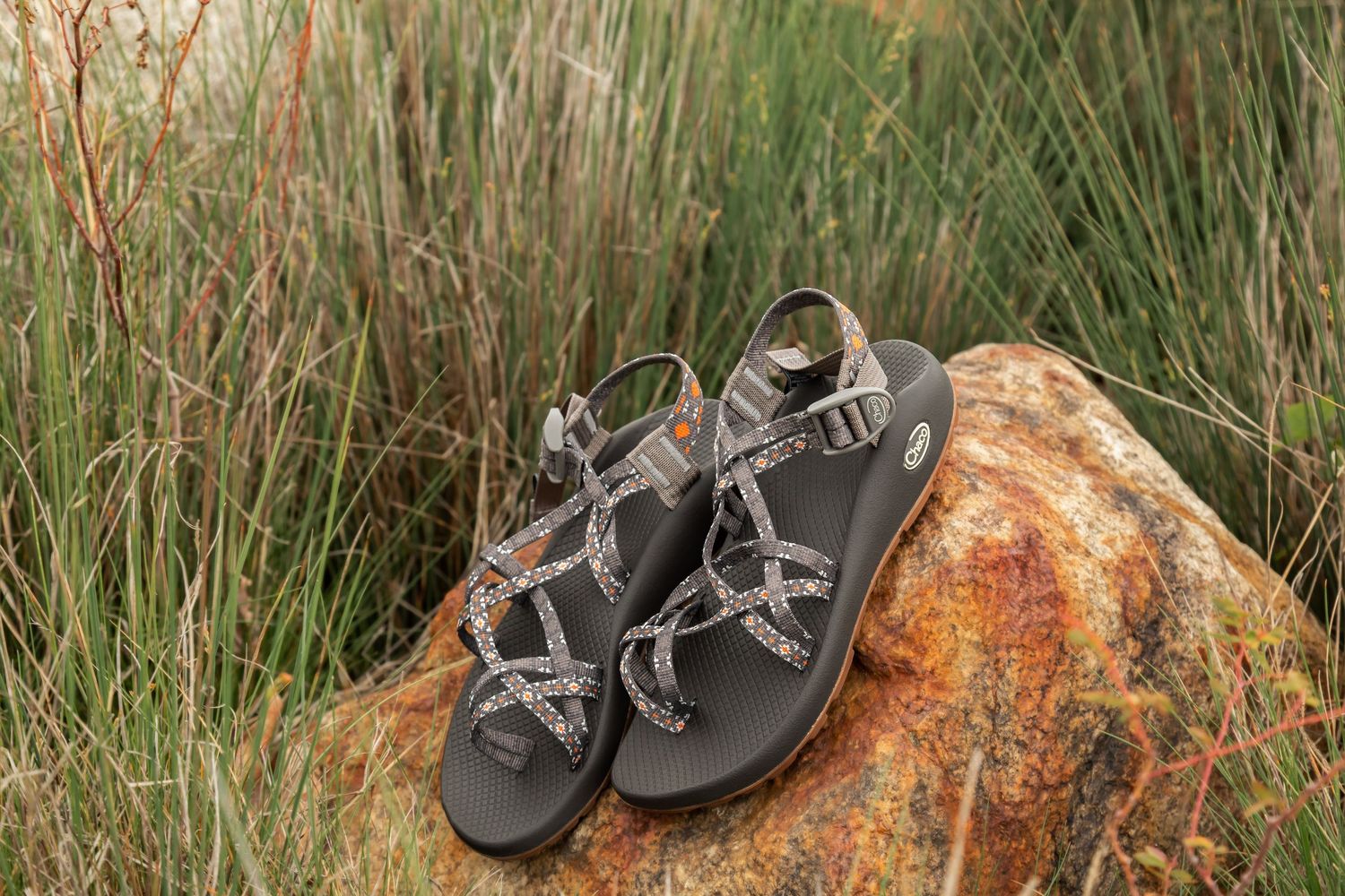 A pair of gray hiking sandals placed on a rustic orange rock surrounded by tall grass.