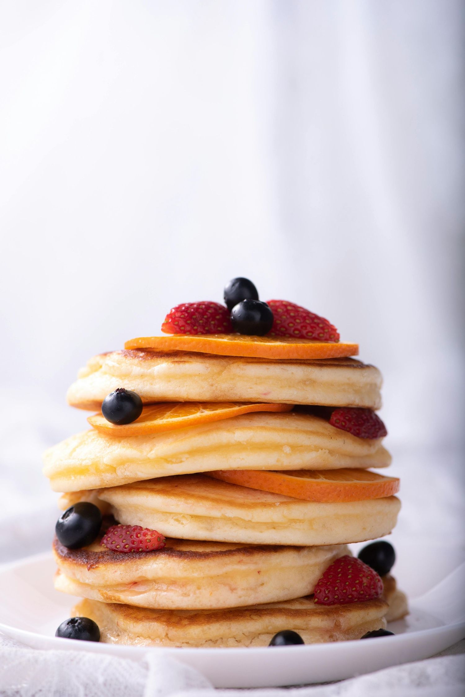 A tall stack of fluffy pancakes topped with fresh strawberries and blueberries on a white plate against light background.