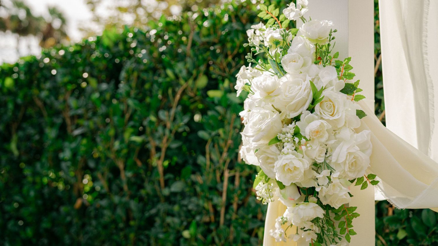 Elegant white floral wedding arch with roses and greenery against a lush garden backdrop.