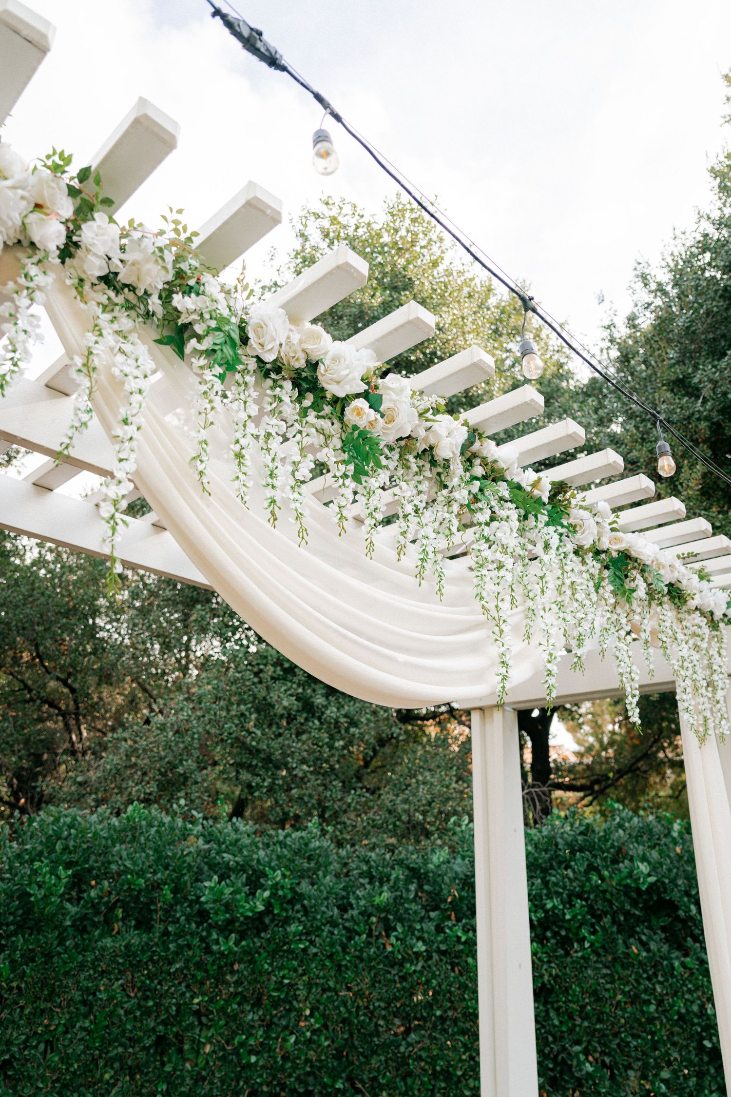 White pergola decorated with flowing fabric drapes and cascading floral vines creates an elegant outdoor wedding setting.