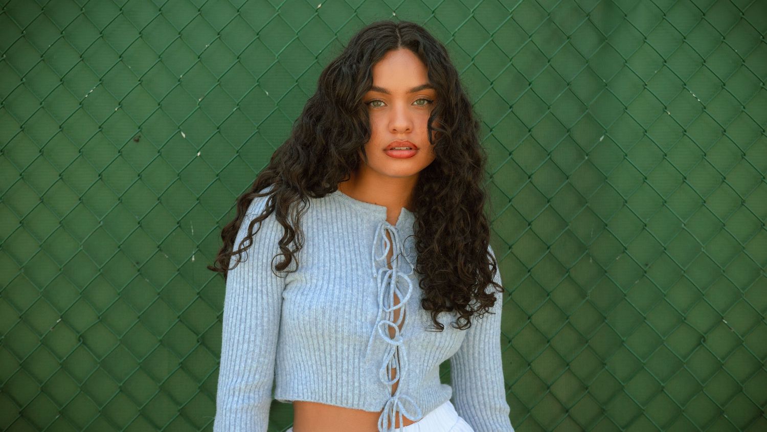 A person with curly dark hair wearing a light blue knit sweater poses against a textured green background.