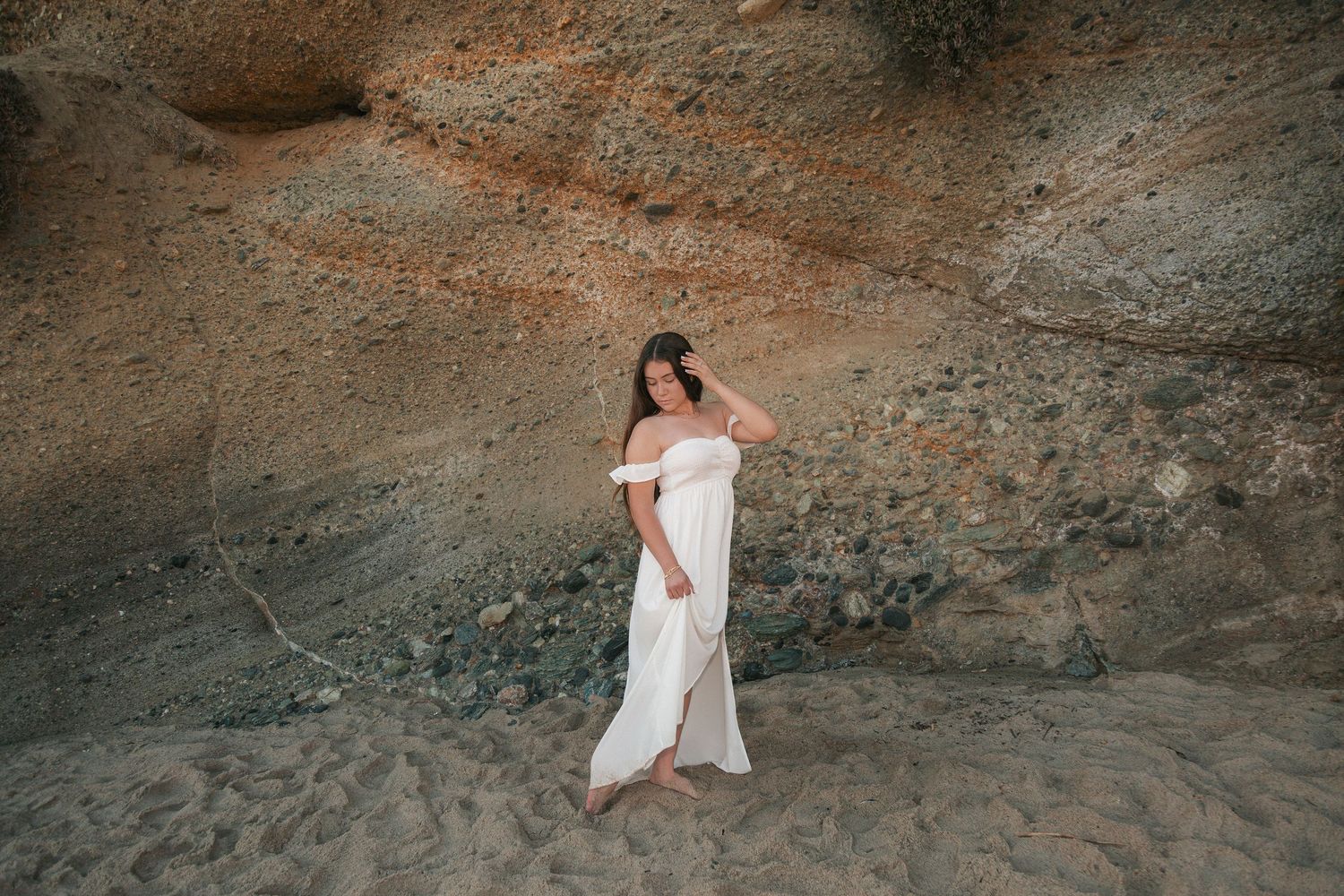A person in a white dress poses gracefully against rugged rock formations during a desert photo session.