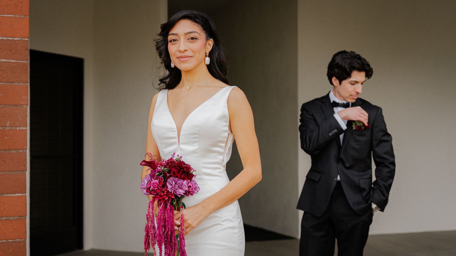 Wedding party members in formal attire pose together in a hallway with white walls and brick accents.