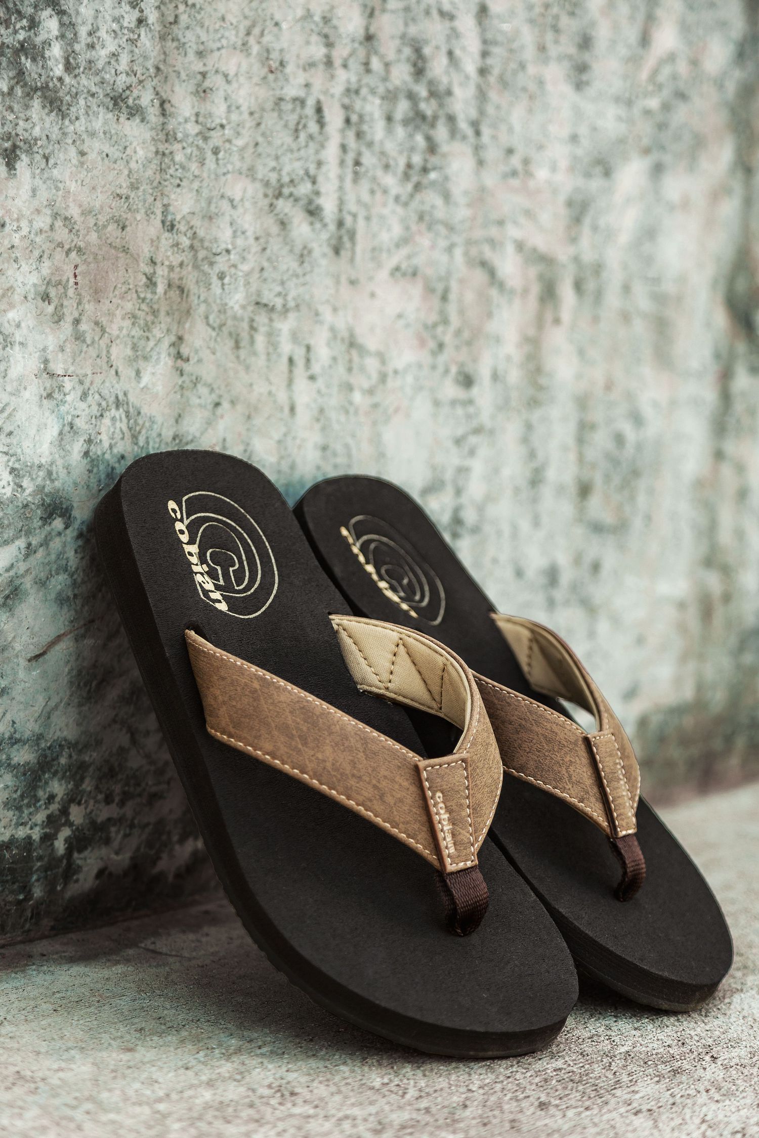 Brown leather flip flops with black soles shown from different angles against a textured concrete background.