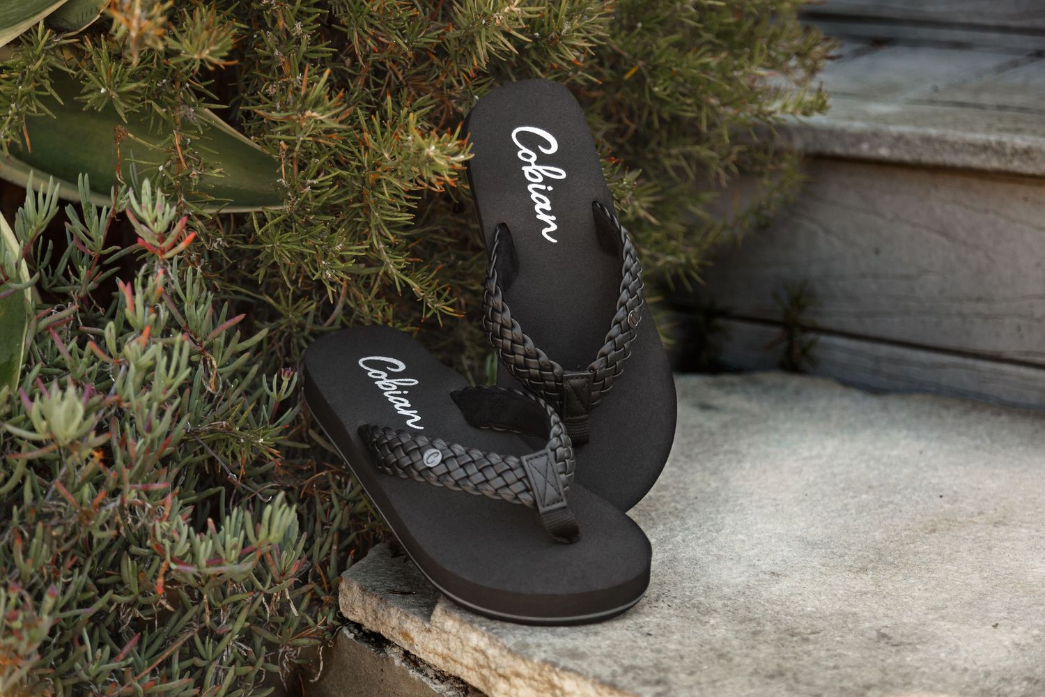A pair of black flip flops with rhinestone embellishments sits on a concrete surface near green plants.