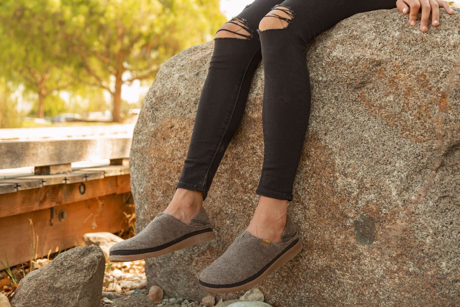 A pair of gray slip-on shoes worn with black leggings while sitting on a large boulder in outdoor sunlight.