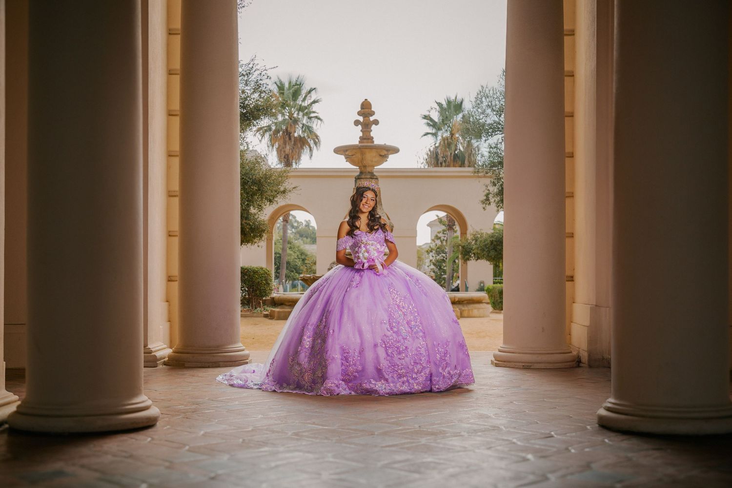 A lavender quinceañera dress sparkles between elegant columns with a fountain and palm trees in the background.