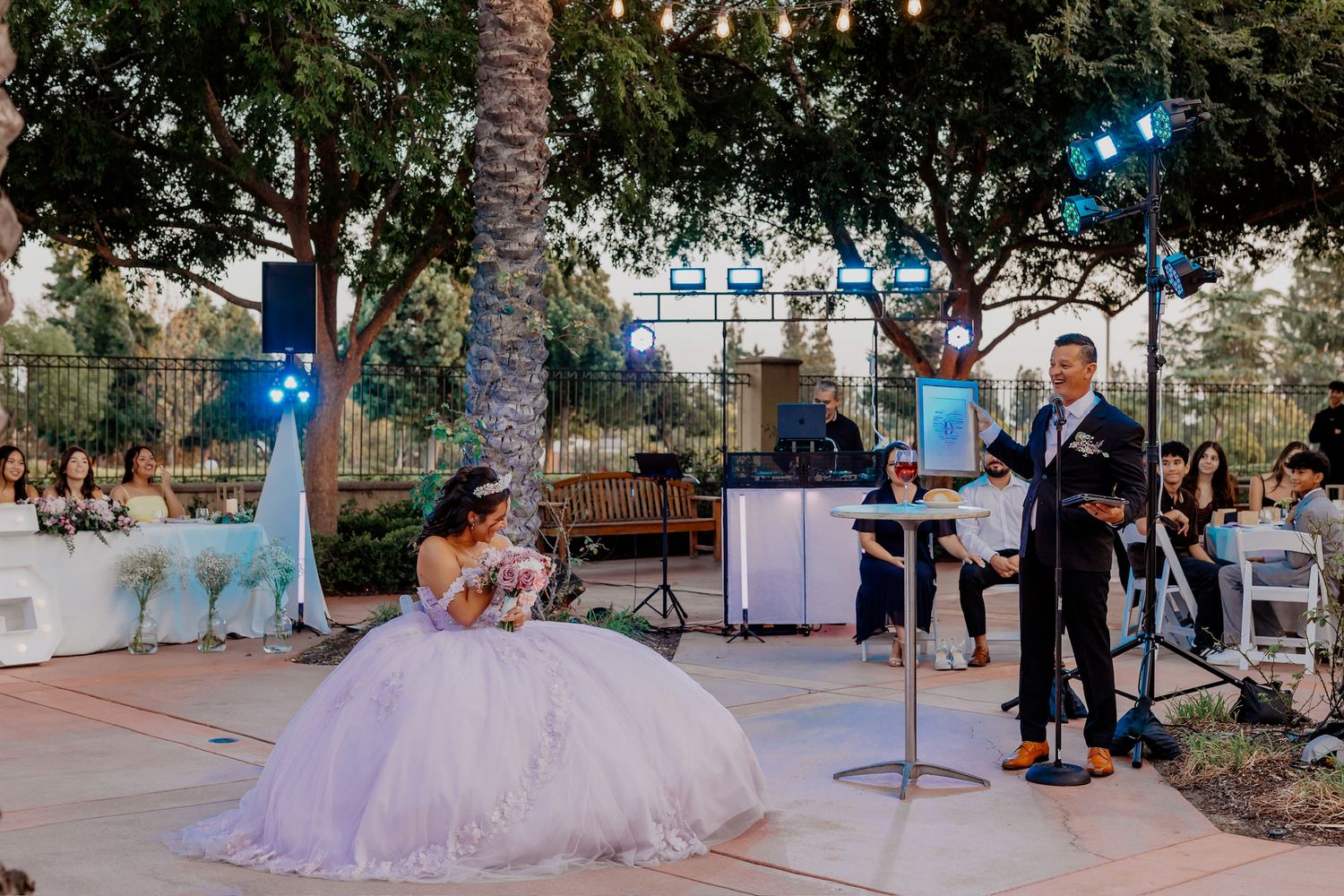 Wedding party poses outdoors at sunset in a garden setting with palm trees and string lights creating a romantic atmosphere.