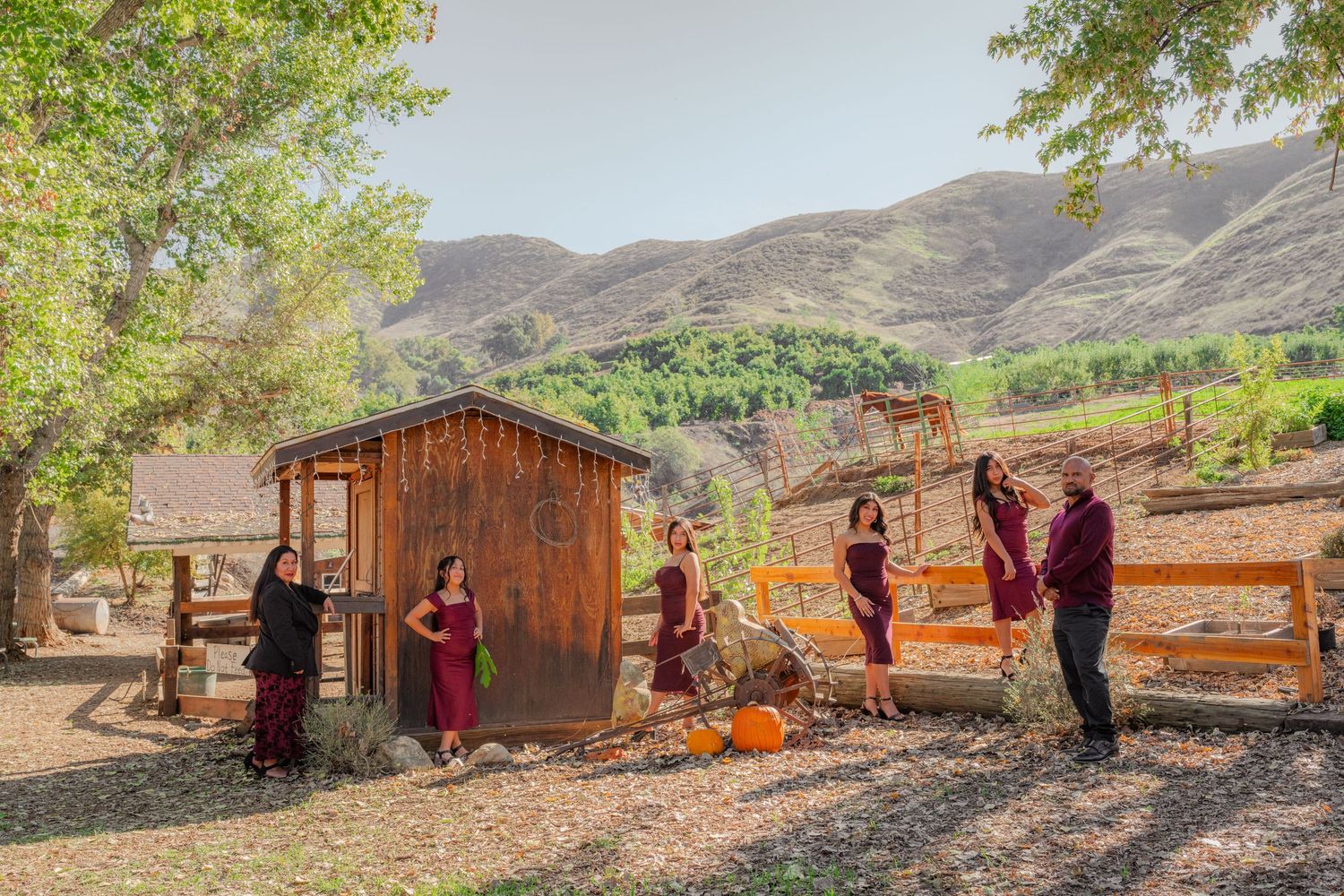 A group of people gather outdoors near a rustic wooden structure with mountains in the background on a sunny day.