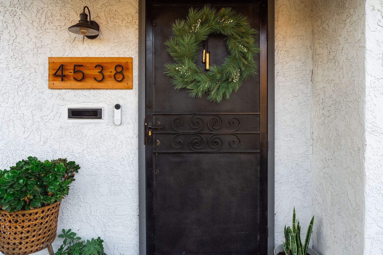 A black front door with a green wreath, orange house numbers 4538, and potted plants on a white stucco exterior.