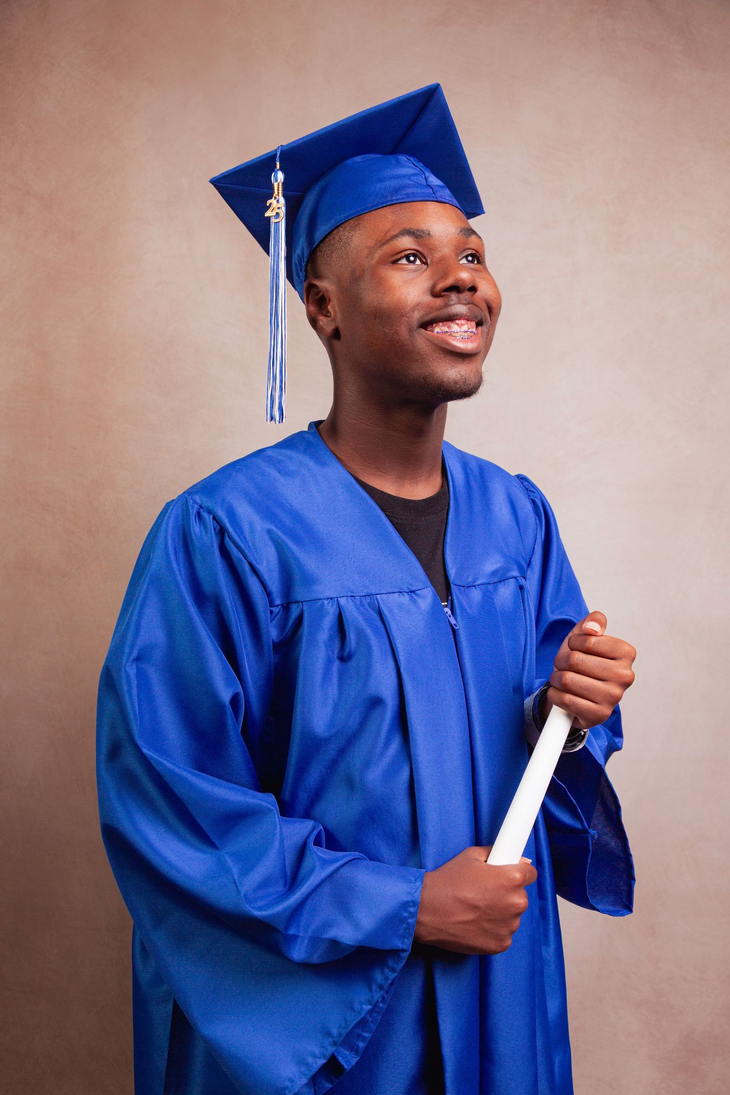 A graduate in bright blue cap and gown holds their diploma while smiling against a neutral backdrop.