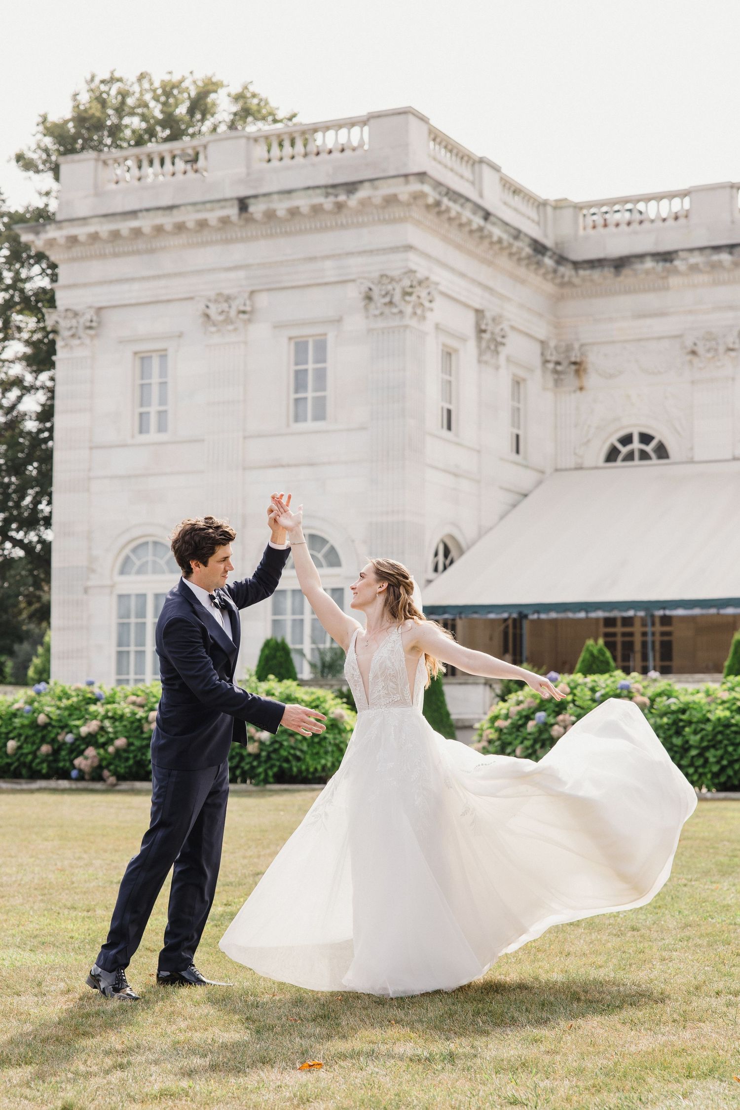 A couple dances joyfully on the lawn in front of an elegant white mansion with classical architecture.