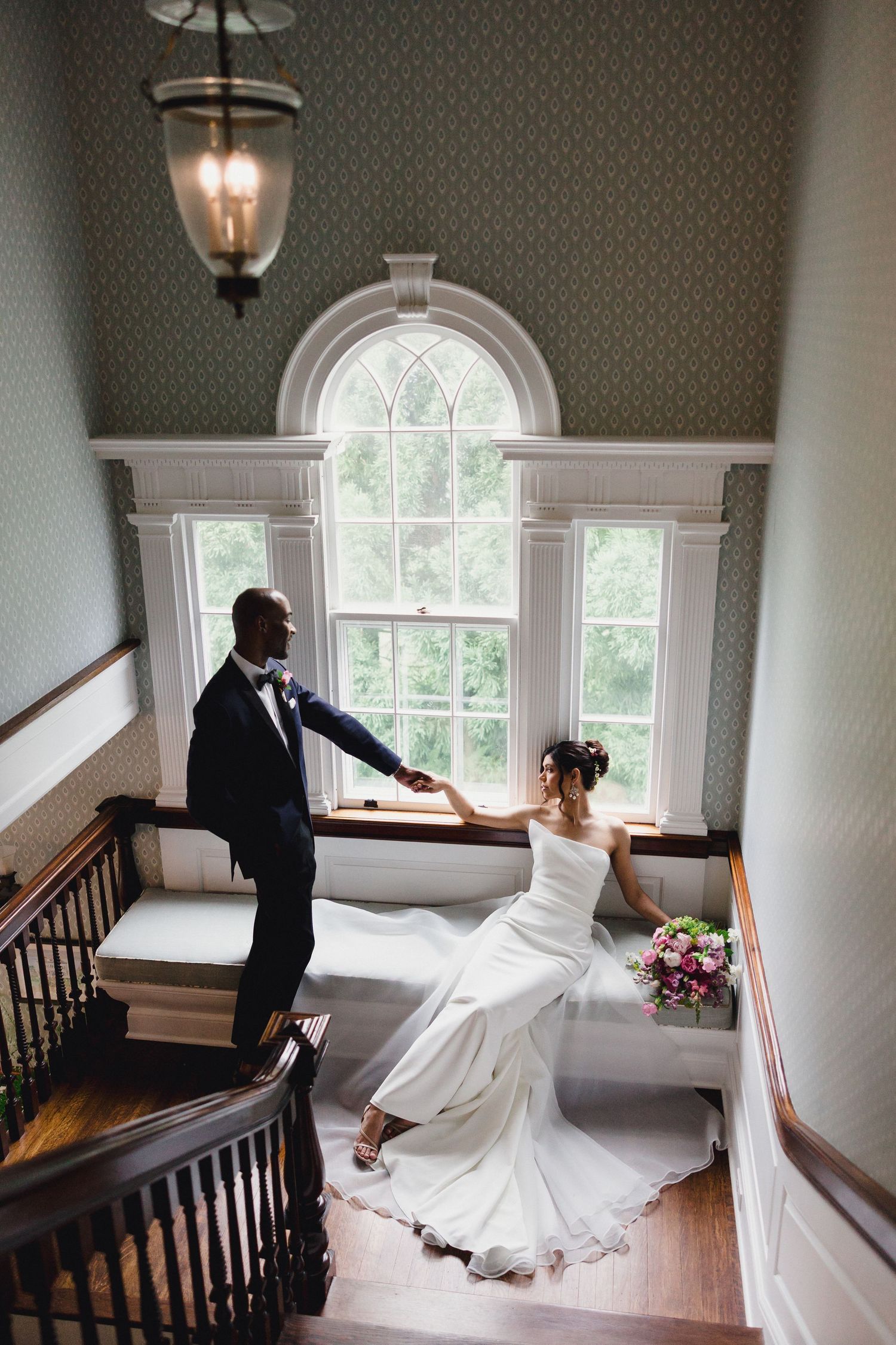 A romantic wedding photo on an elegant staircase with an arched window and flowing white dress against dark wood details.