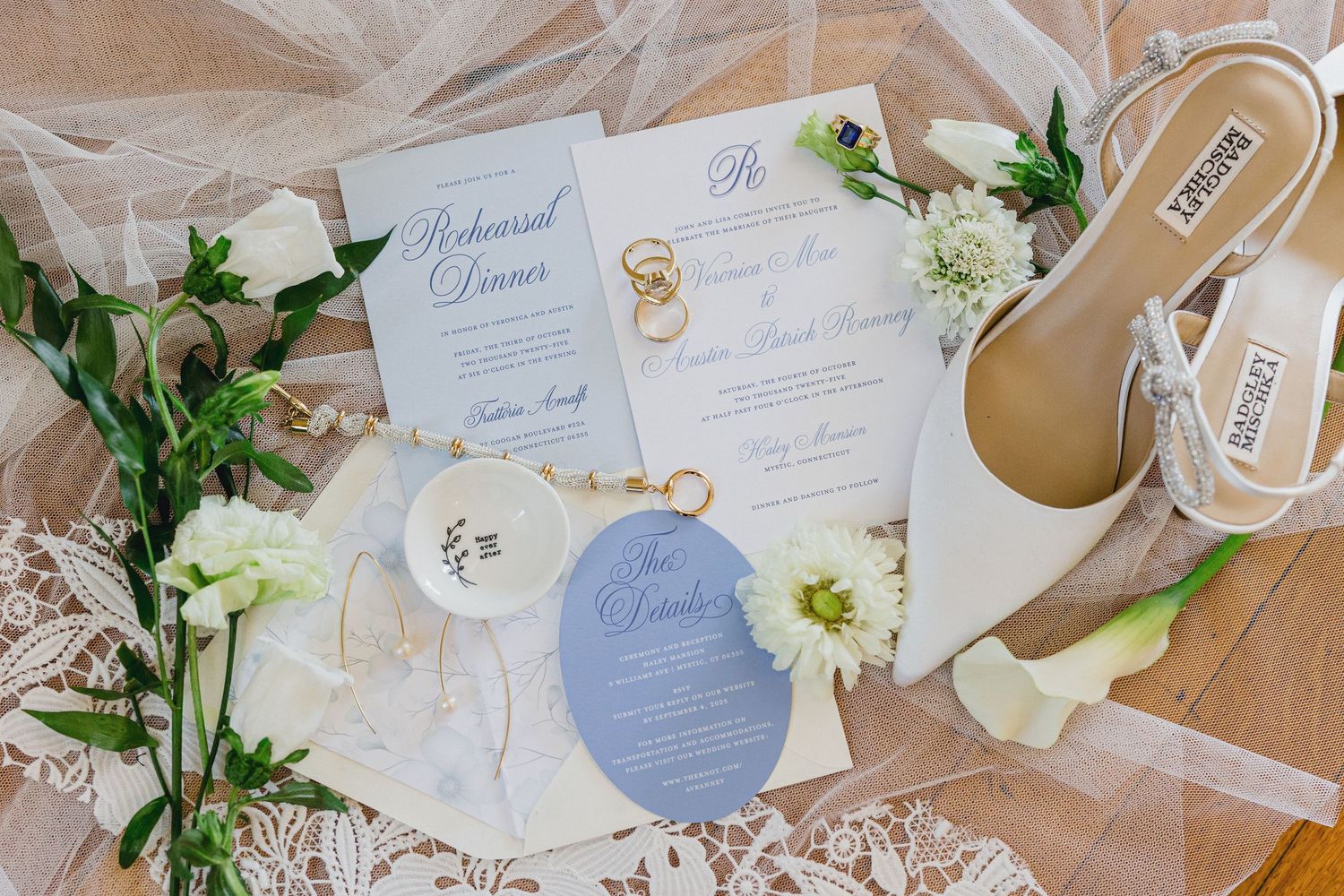 Elegant wedding invitation suite with white floral arrangements, blue accents, and delicate ribbons laid out on wooden surface.