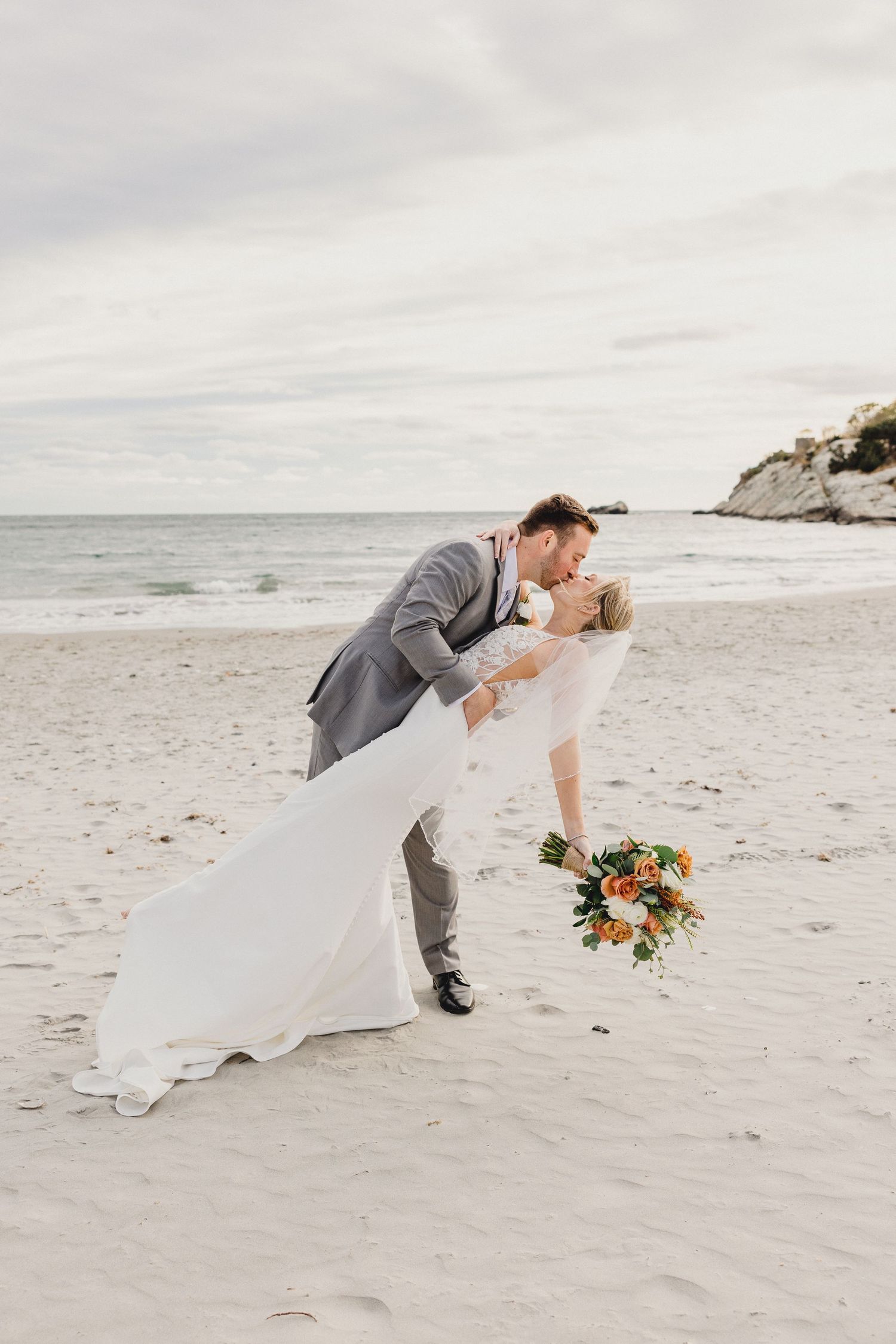 A romantic beach wedding scene with a couple embracing in a dip kiss while holding a vibrant orange and white bouquet.