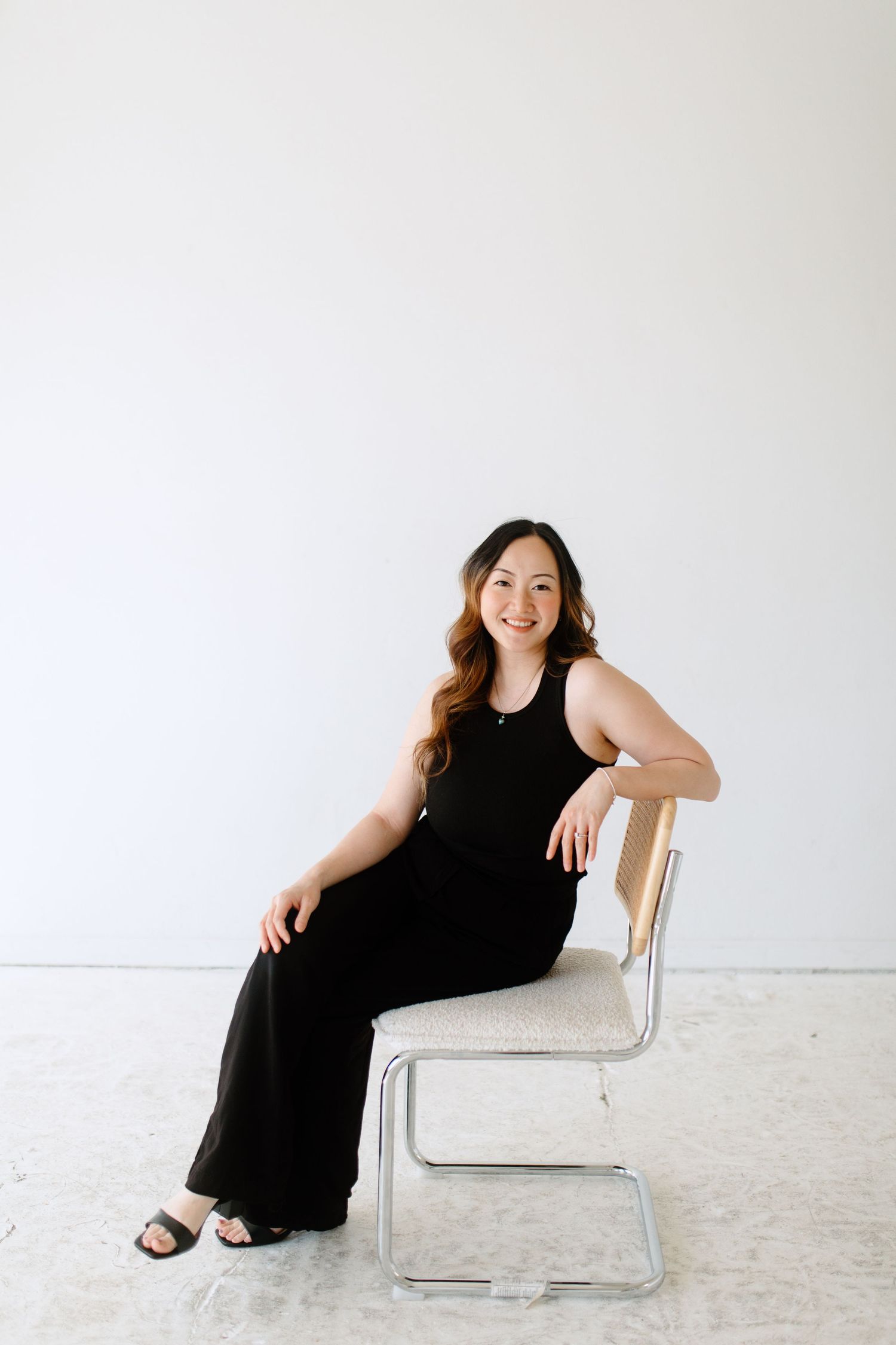 A figure in a black dress poses gracefully on a modern white chair against a minimalist white studio backdrop.