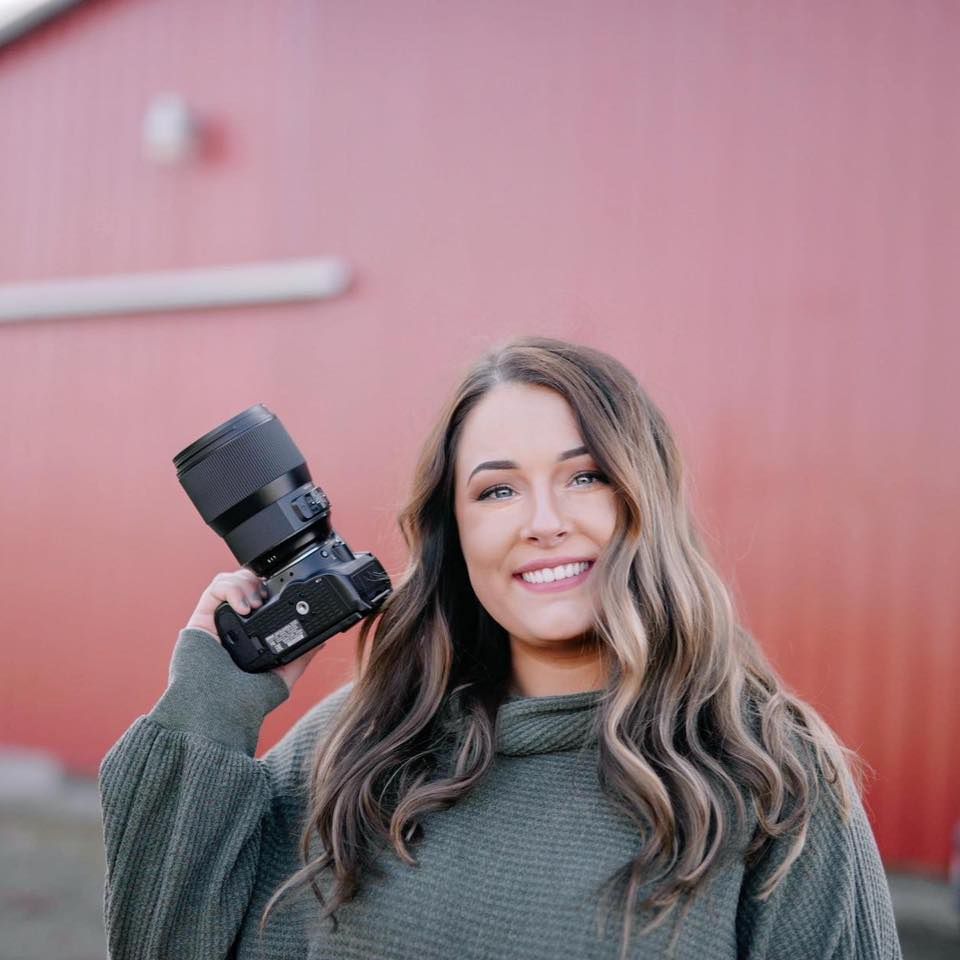 Professional photographer wearing a gray sweater holds up a camera against a pink wall background.