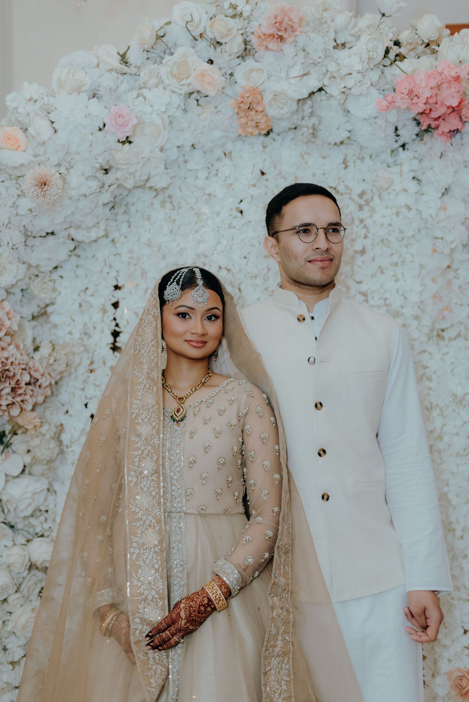 A traditional wedding couple poses against a floral backdrop of white and pink roses in elegant attire.