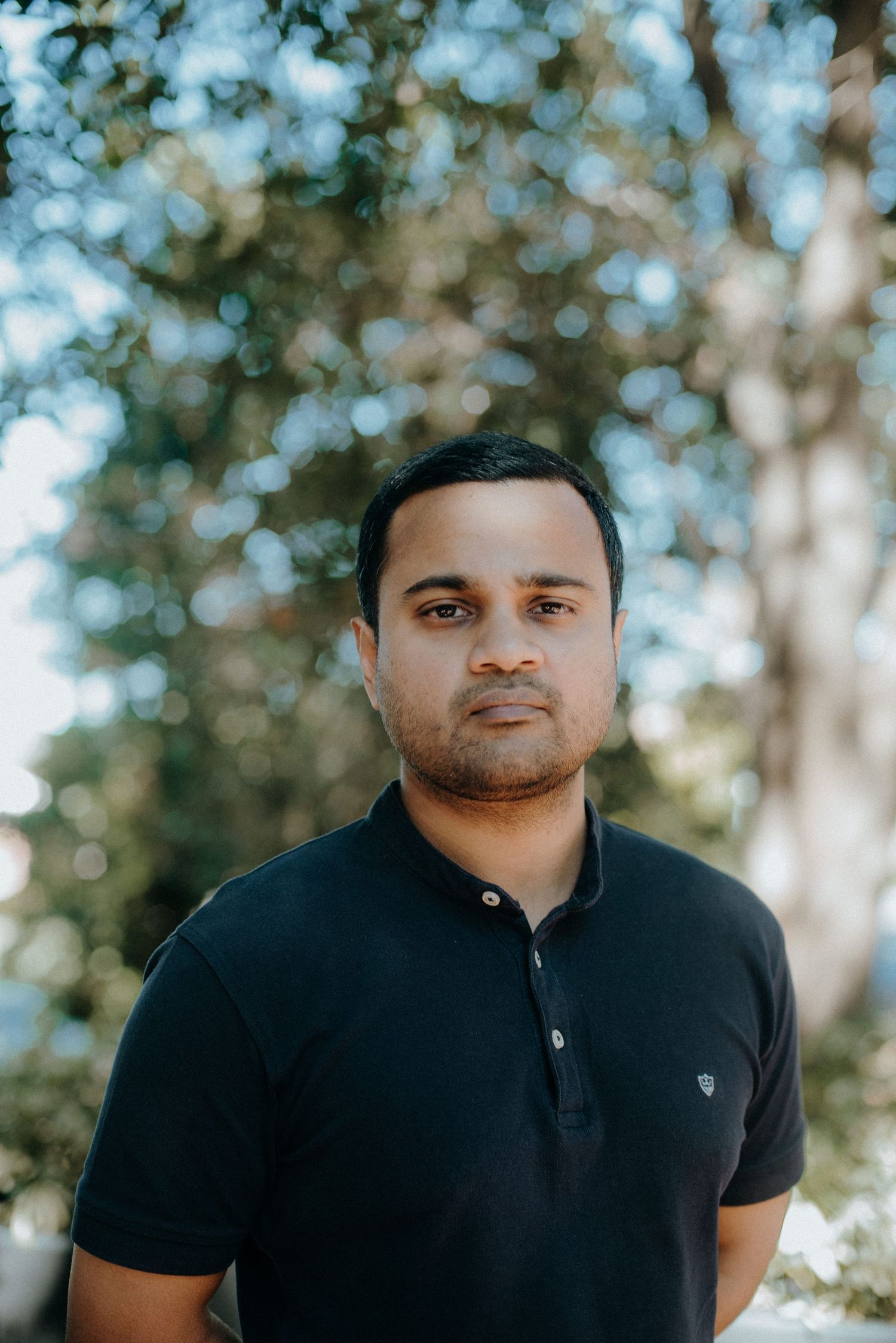 Person stands outdoors wearing a black polo shirt against a blurred natural background with bokeh lighting effect.