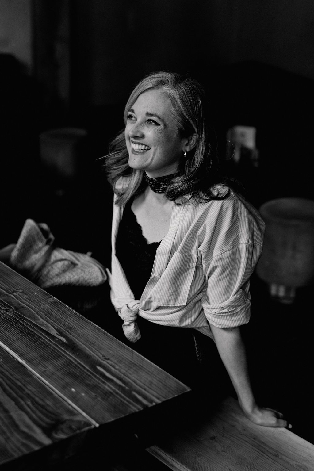 Black and white photo showing a person leaning on a wooden table while smiling and wearing a white button-up shirt.