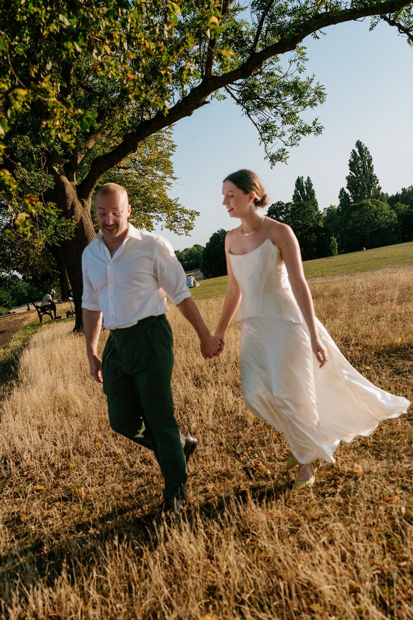 A joyful couple holds hands while walking through a golden field, the bride in a flowing white dress and groom in white shirt.
