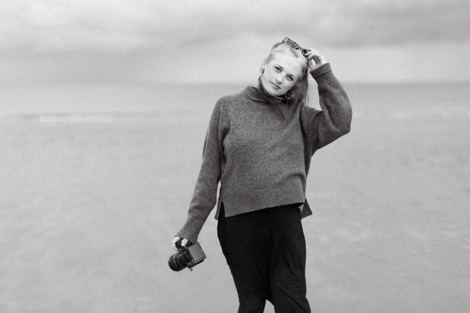 Black and white photo of a woman holding a camera on a beach, wearing a turtleneck sweater on a cloudy day.