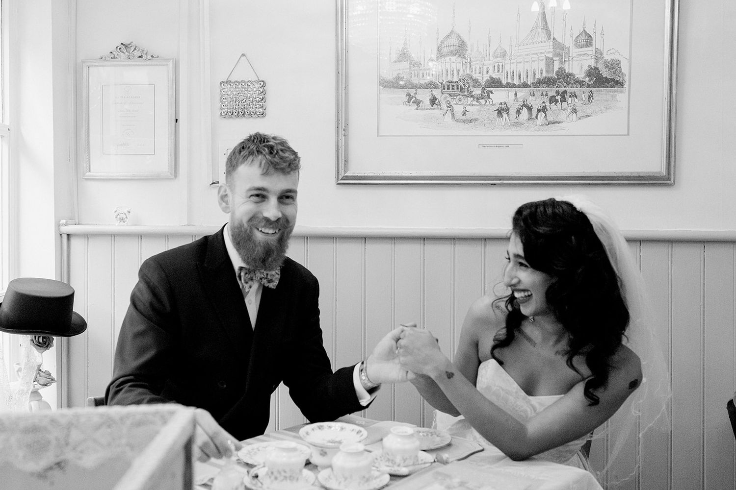 Black and white photo of a smiling bride and groom sitting together at a table with tea cups at their wedding reception.