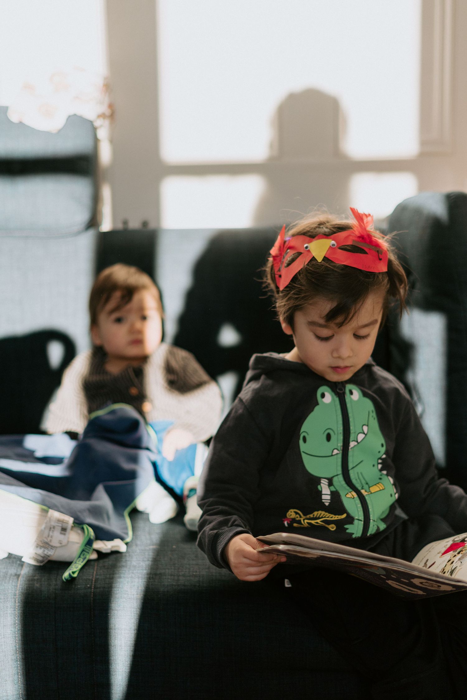 Young child wearing dinosaur sweatshirt and red bow reads quietly on dark couch in soft indoor lighting.