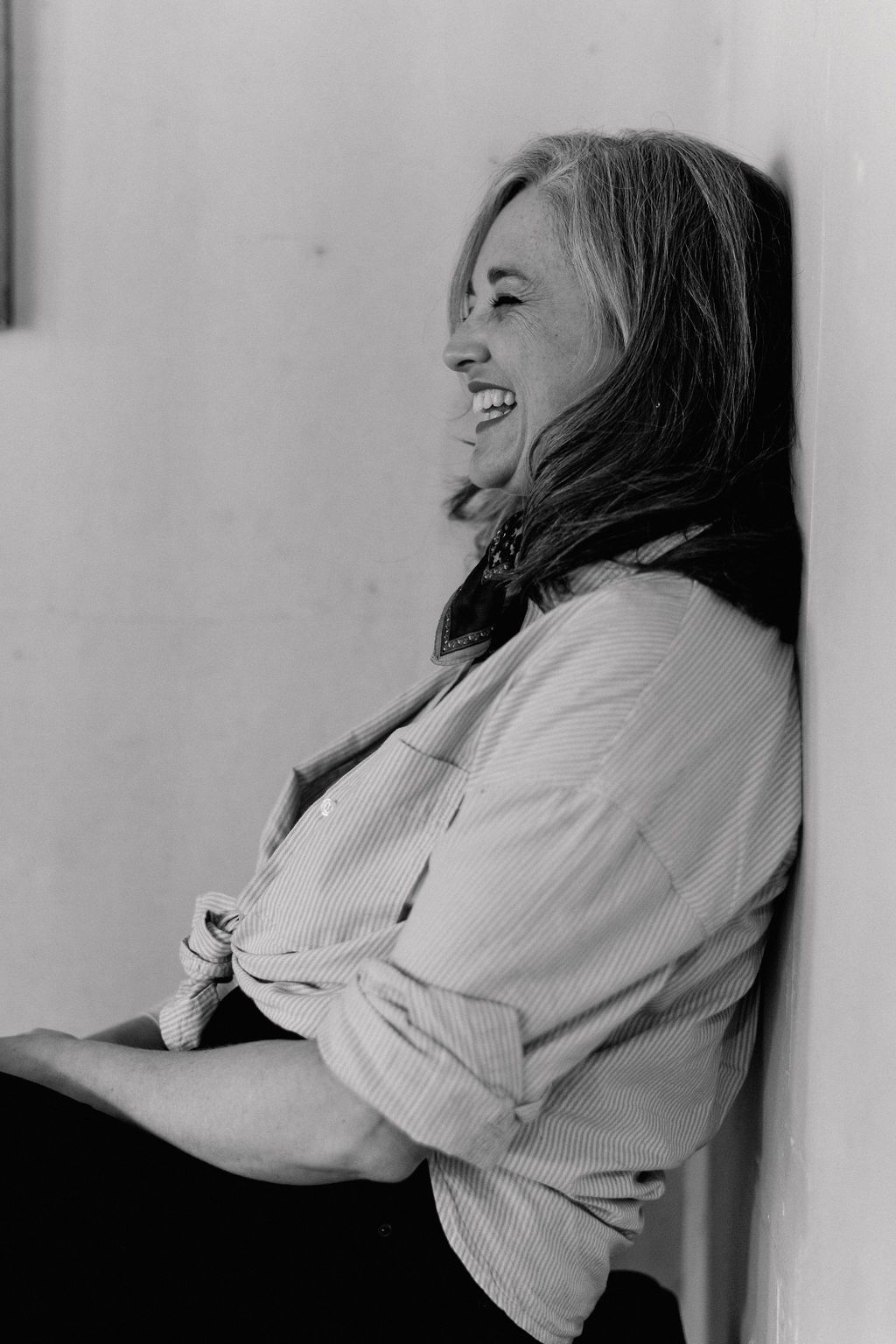 Black and white portrait of a person laughing joyfully while leaning against a wall wearing a light colored button-down shirt.