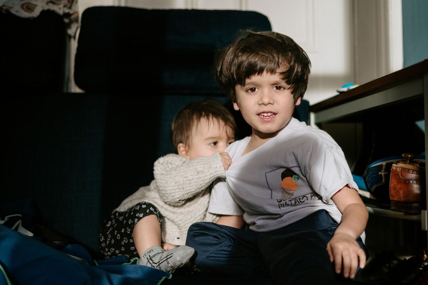 Two children sit together on a couch, with one wearing a white t-shirt with a logo and the other in casual attire.