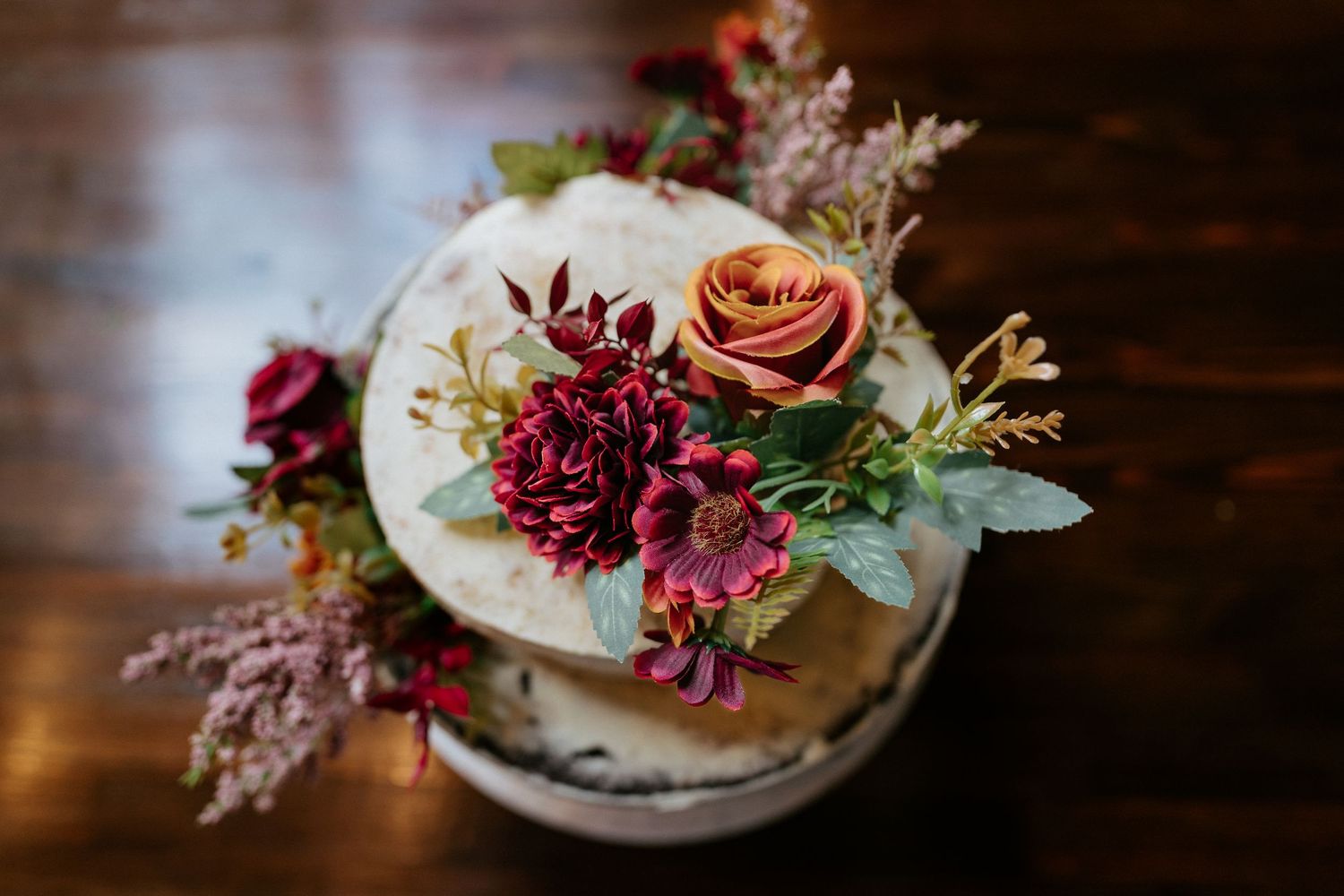 Rustic naked wedding cake topped with burgundy dahlias, peach roses, and greenery on a dark wood table.