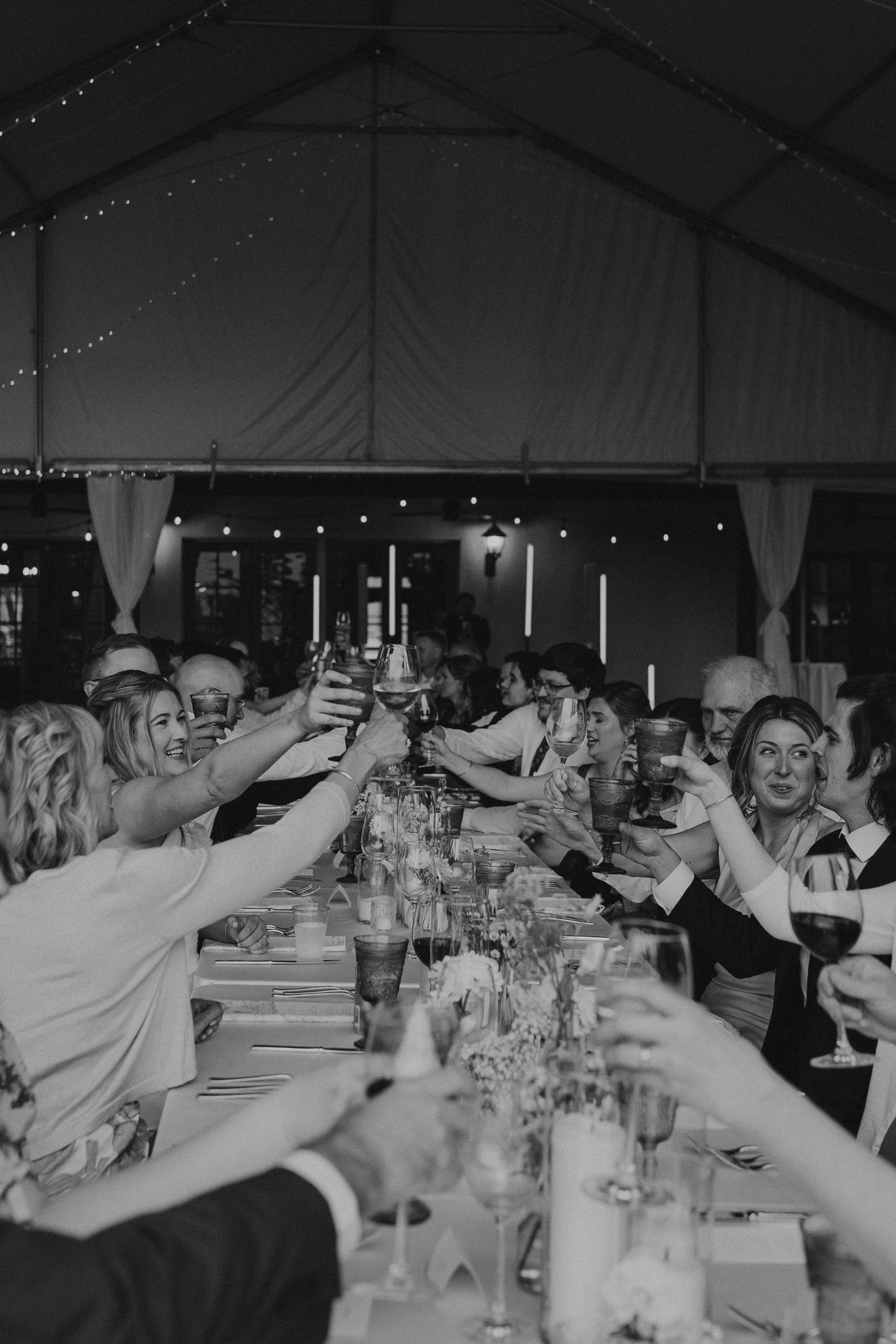 Black and white photo of wedding guests raising glasses in a toast at a long reception table under a marquee tent.