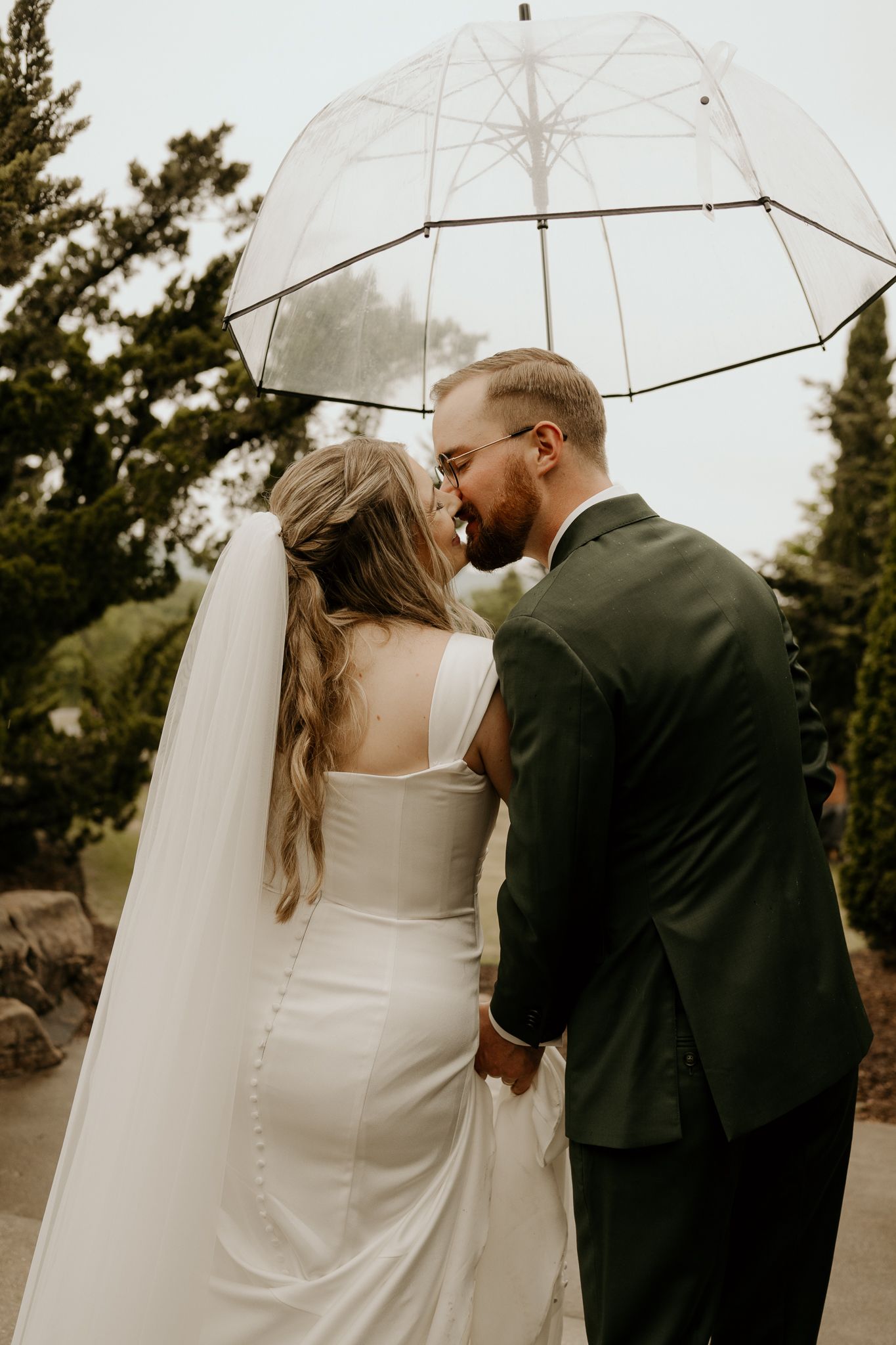 Bride and groom share a romantic moment under a clear umbrella on their rainy wedding day, holding hands outdoors.