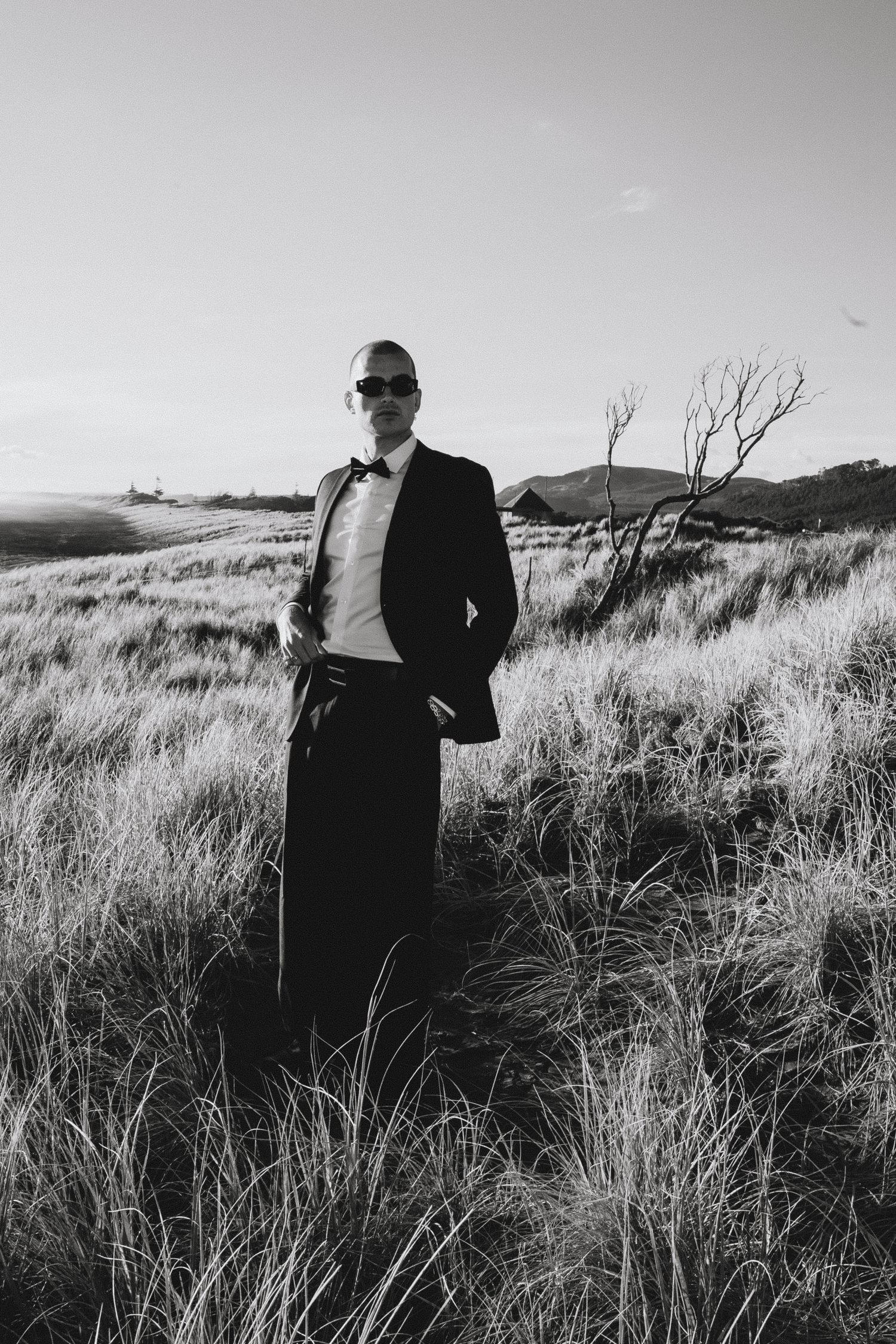 A black and white portrait of someone in formal attire standing in tall grass against a moody landscape.
