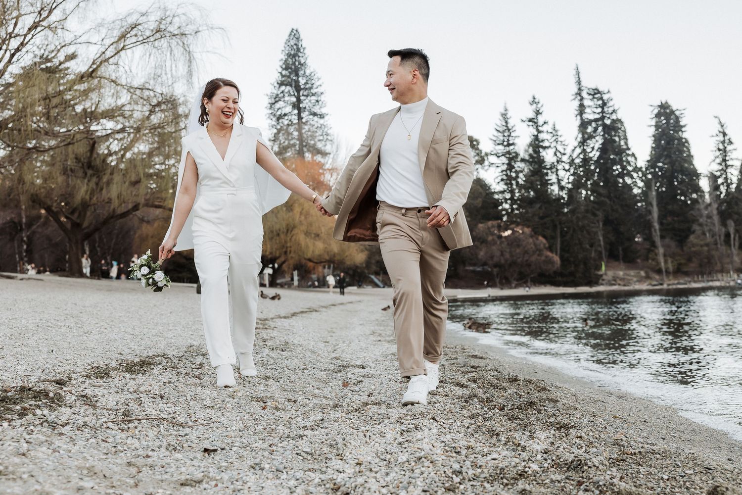 A couple in wedding attire walks hand in hand along a snowy winter path near a lake and evergreen trees.