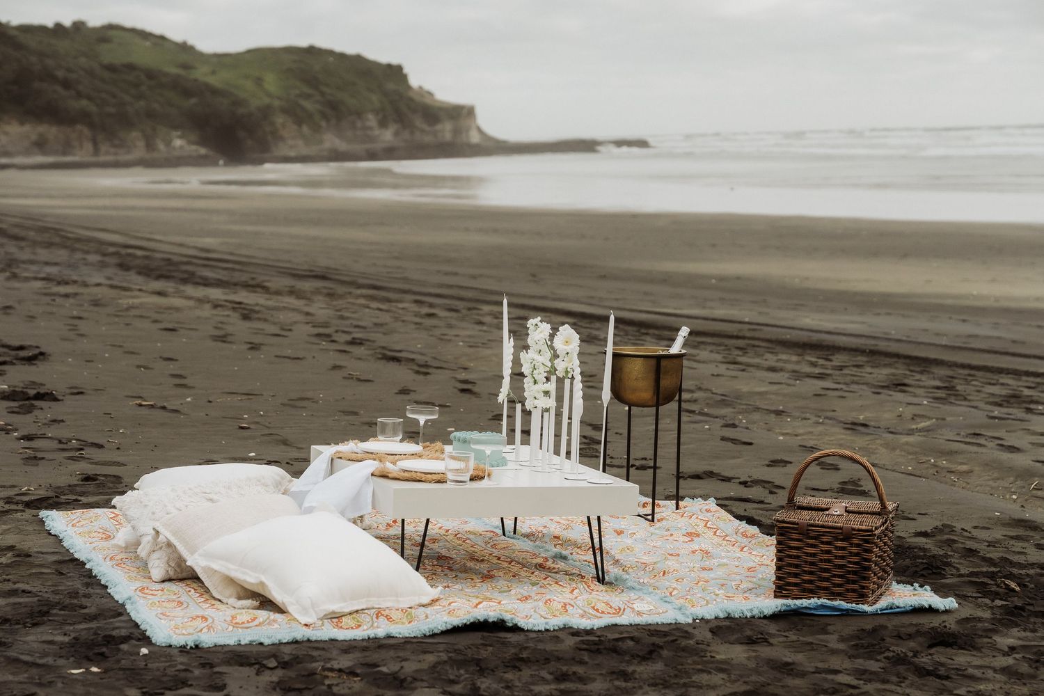 A rustic beach picnic setup with white linens and wicker baskets on a sandy shore near coastal cliffs.