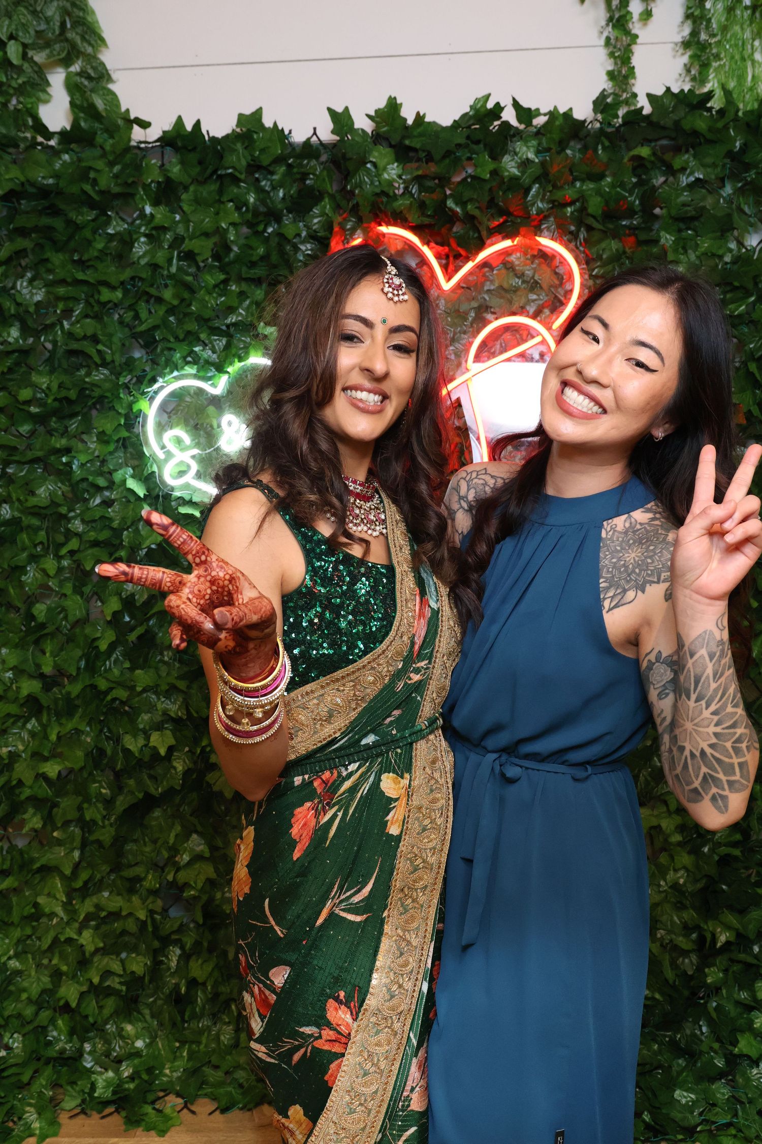 Two friends in traditional and modern attire pose playfully against an ivy-covered wall with neon light effects.