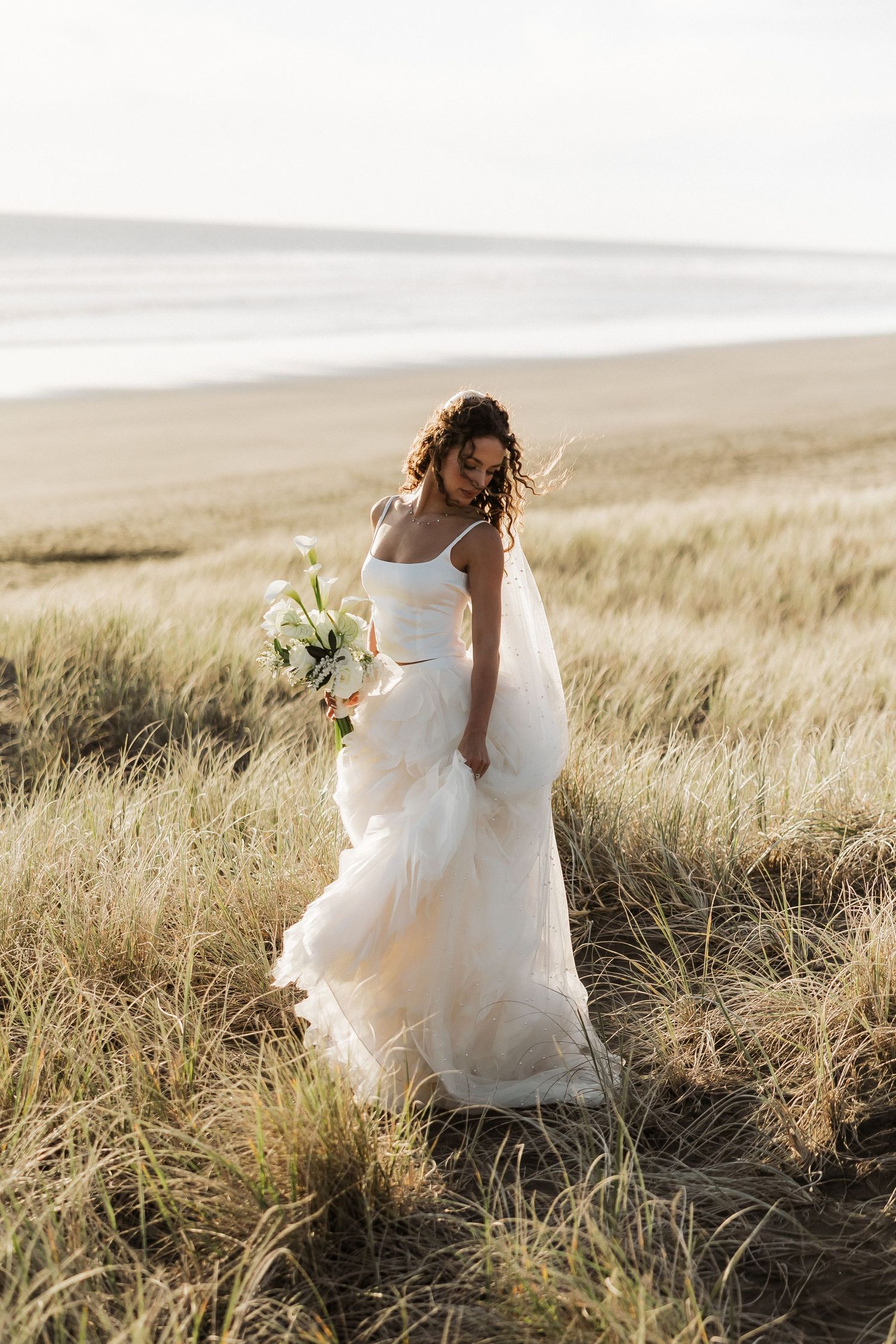 Bride in flowing white dress walks through sunlit field of tall grass.